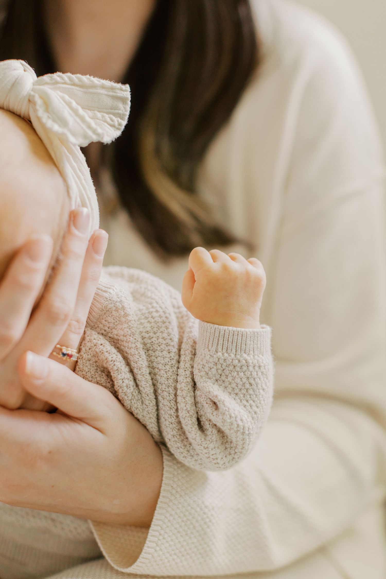 A mother in a cream sweater tenderly holds her newborn baby dressed in a knit outfit with a bow headband.
