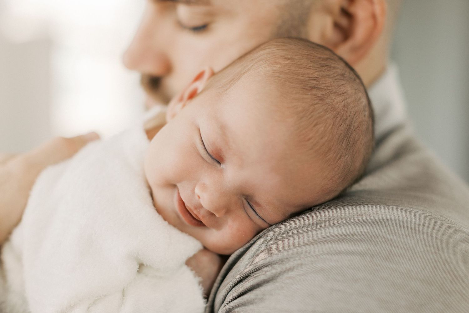 Peaceful newborn baby sleeping on father's shoulder, wrapped in white blanket in a tender bonding moment.