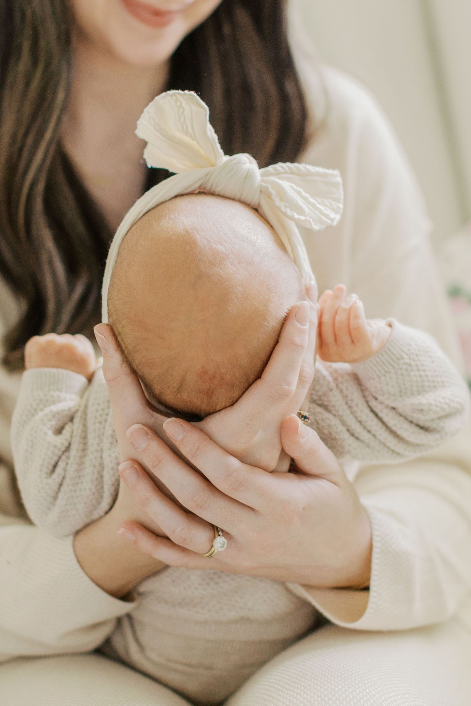 Mother gently cradling newborn baby girl wearing a white bow headband, close-up portrait photo.