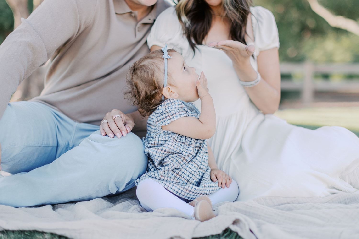 A family sits on a blanket outdoors, with a toddler in a gingham outfit between two adults in soft neutral tones.