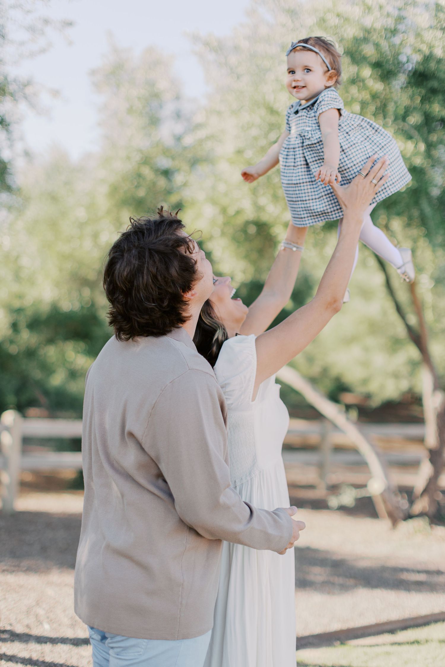 A couple joyfully lifts their smiling toddler in the air outdoors, surrounded by lush green trees.