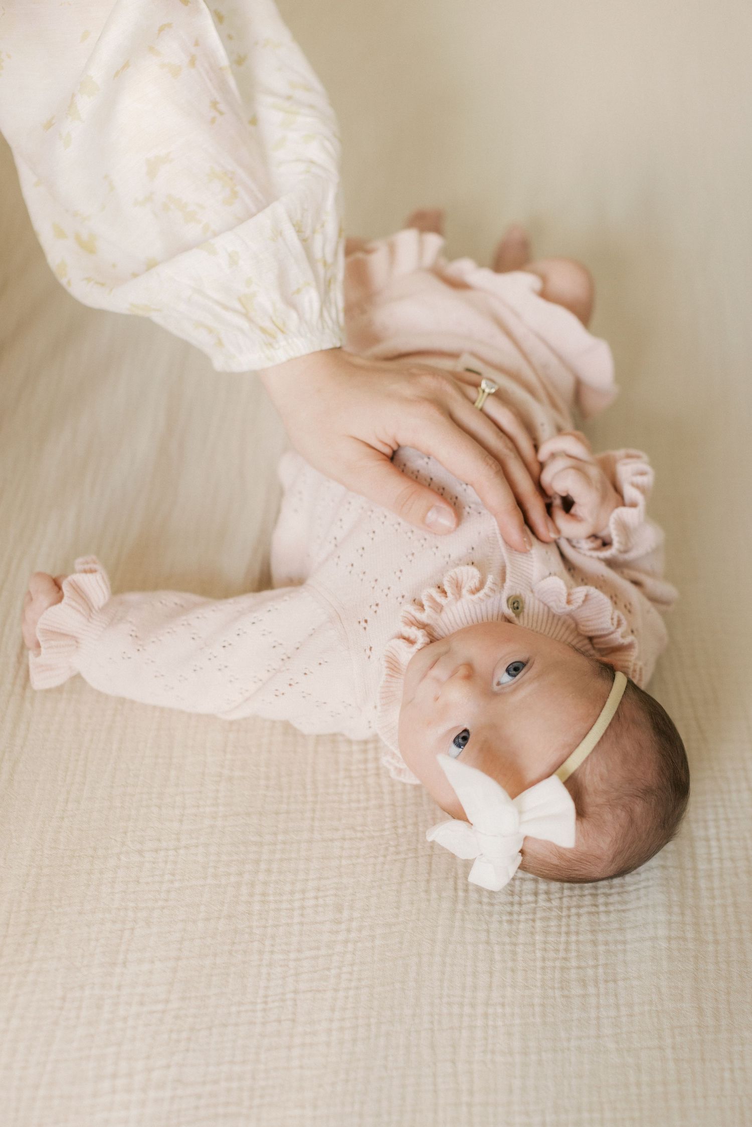 Newborn baby girl in pink ruffle outfit and white bow headband, gently held by mother's hand on soft blanket.