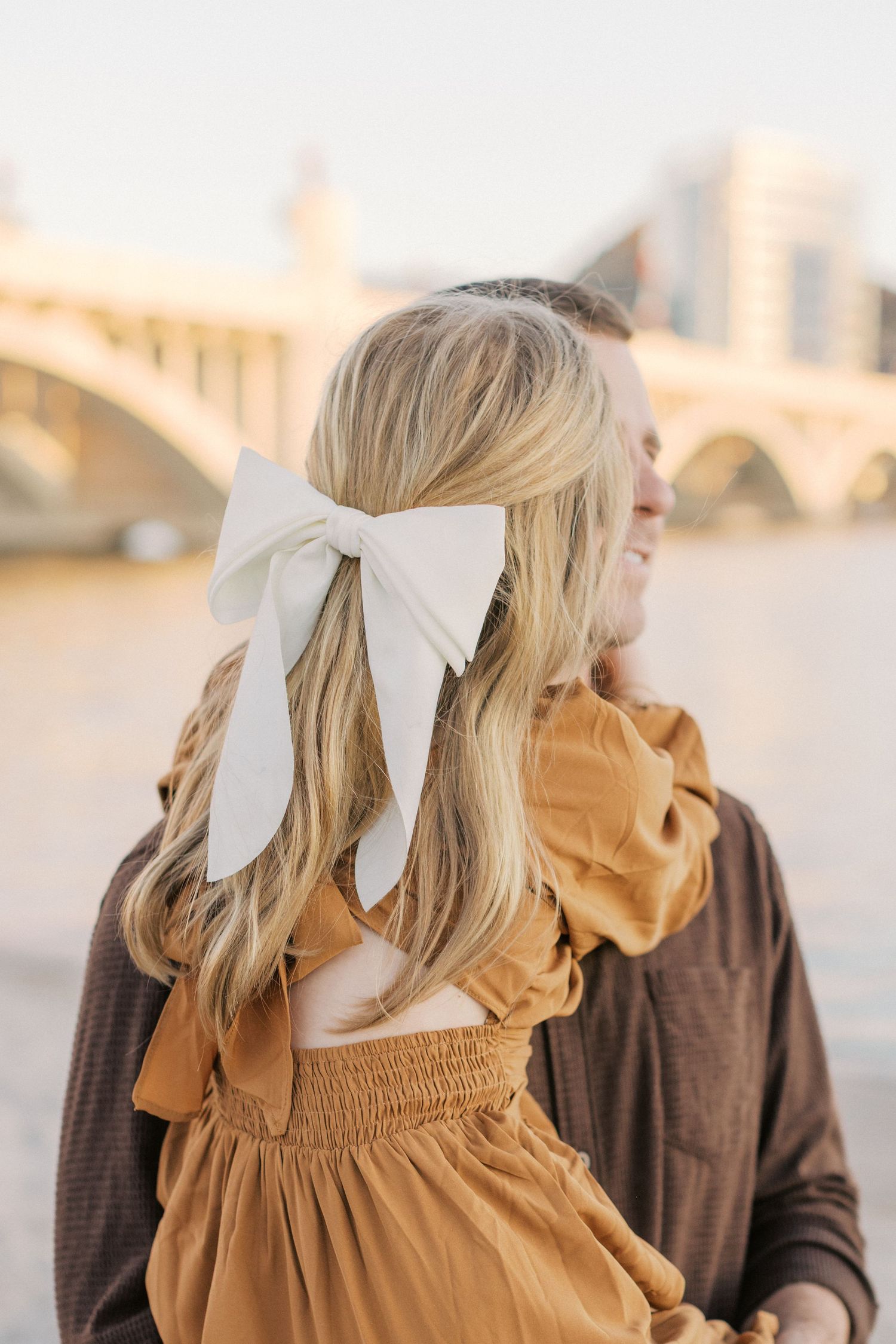 Blonde woman with white bow hair accessory in mustard dress stands near a bridge with a smiling man in brown shirt.