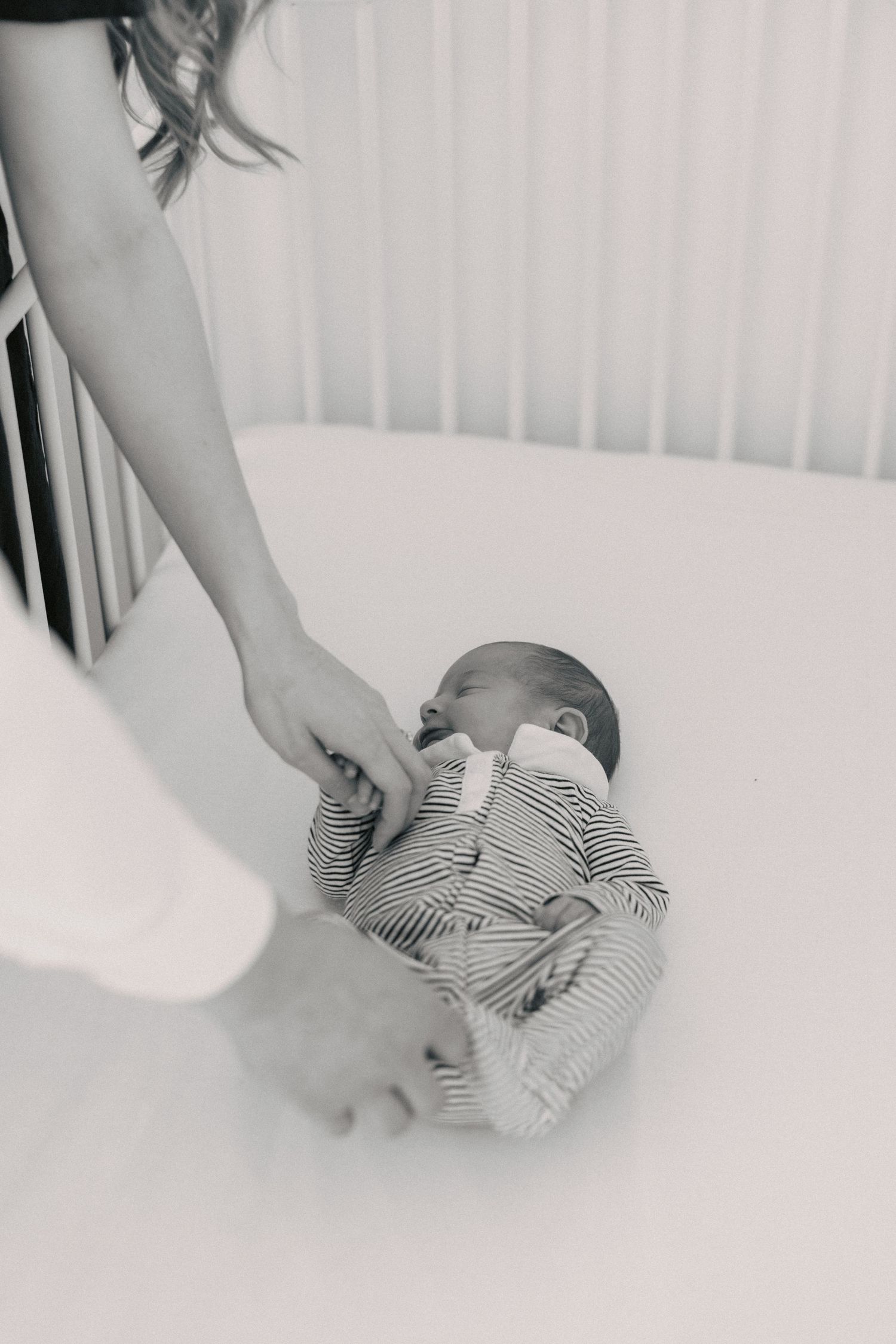 Black and white photo of a newborn baby in a striped onesie lying in a white crib while a parent gently holds their hand.