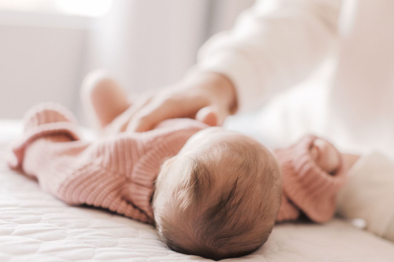 Newborn baby in pink knit outfit lying on white surface while parent gently touches them.