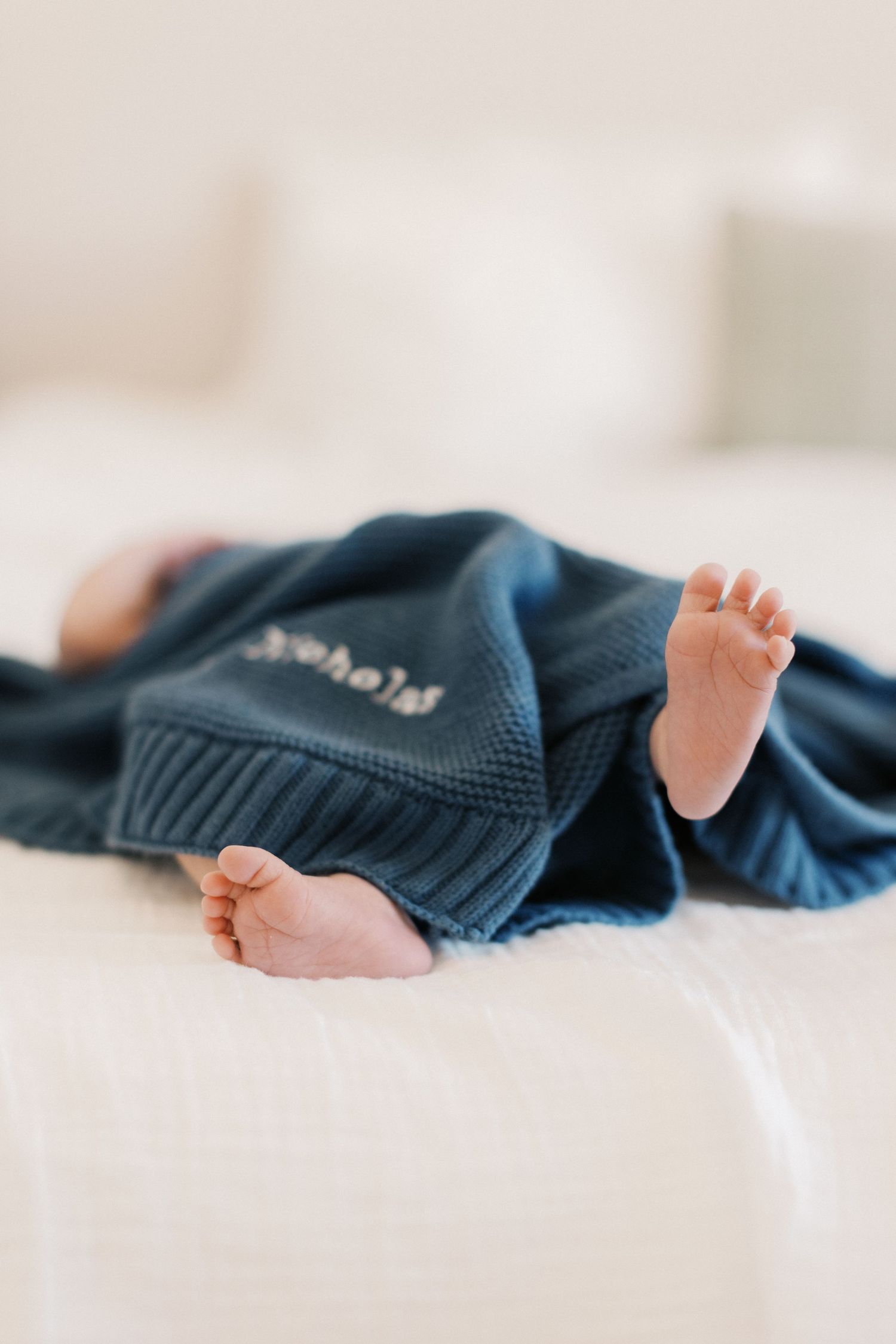 Newborn baby feet peeking out from a navy blue knit personalized blanket on a white bed.