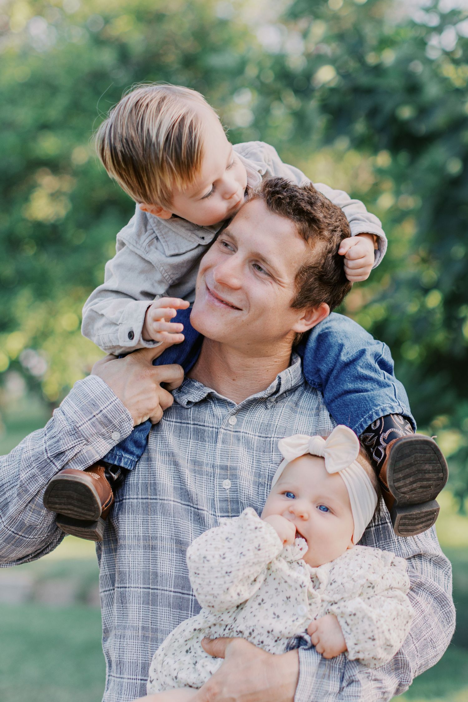 Smiling father holds baby girl while toddler son sits on his shoulders outdoors in green park setting.