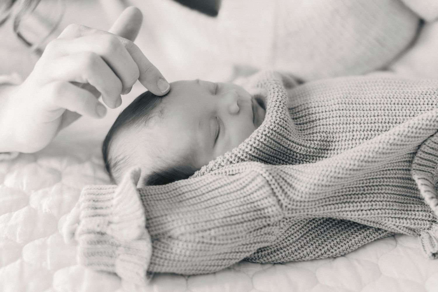 Black and white close-up of sleeping newborn baby wrapped in knit blanket with parent's hand.