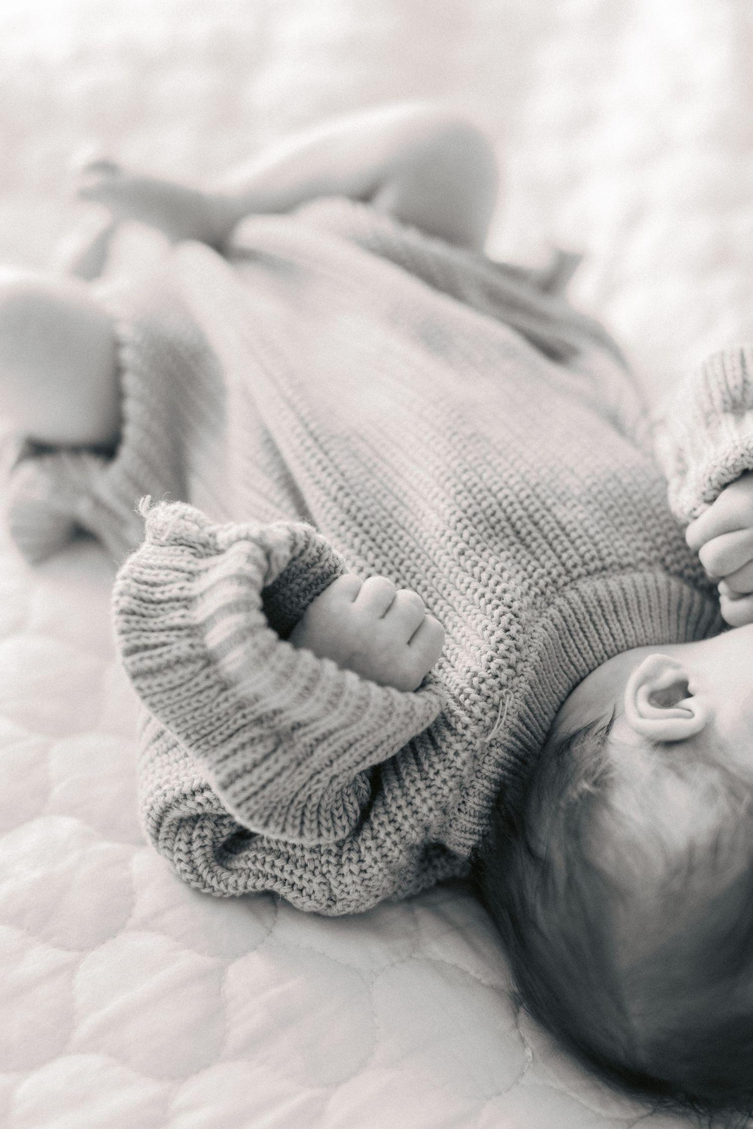Black and white photo of a newborn baby in a knit romper lying on a quilted white blanket.