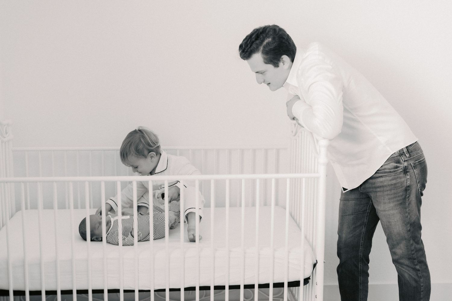 Father leaning over white crib looking at baby in black and white nursery photo.