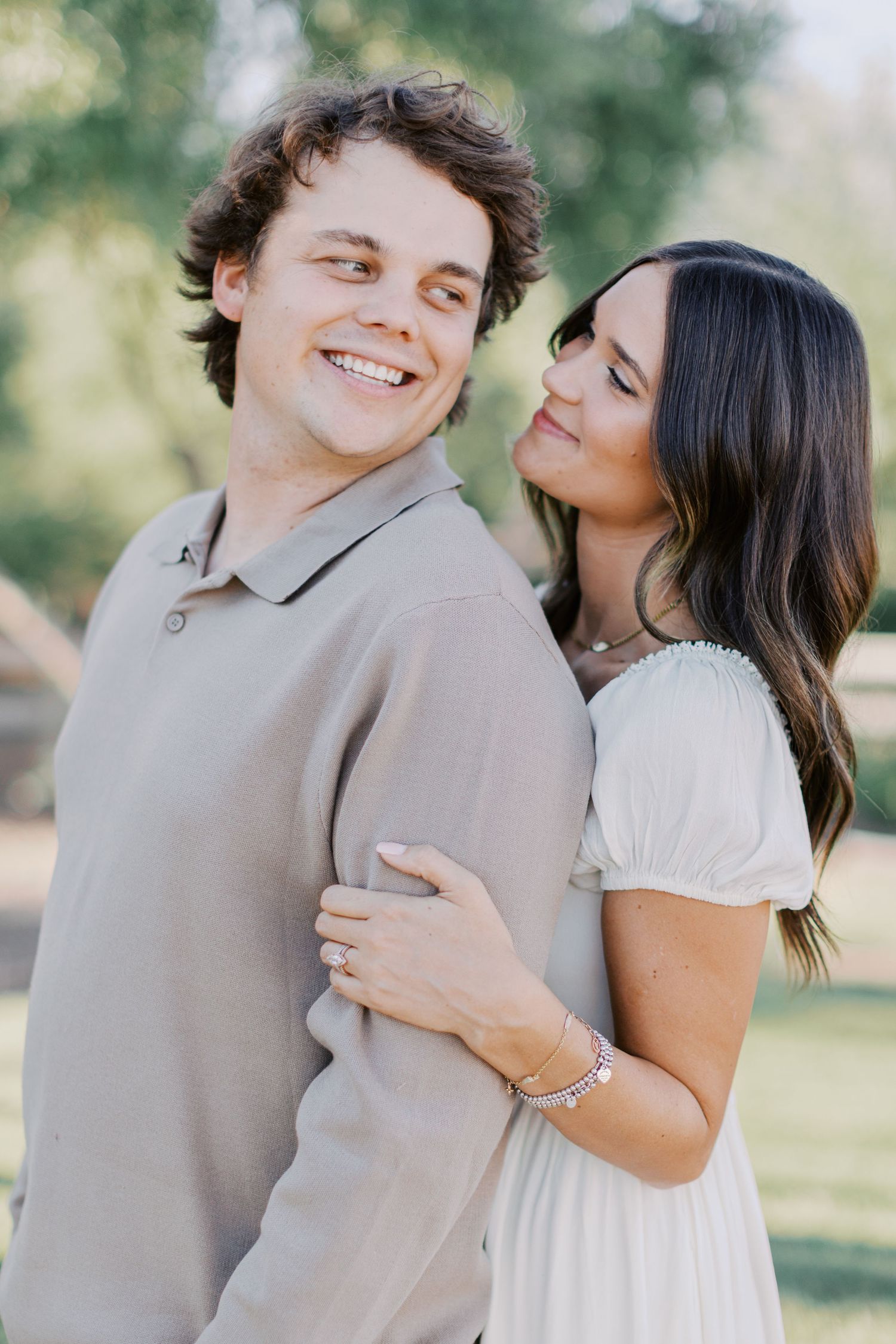 A smiling couple poses outdoors, man in beige polo, woman in white dress embracing him from behind.