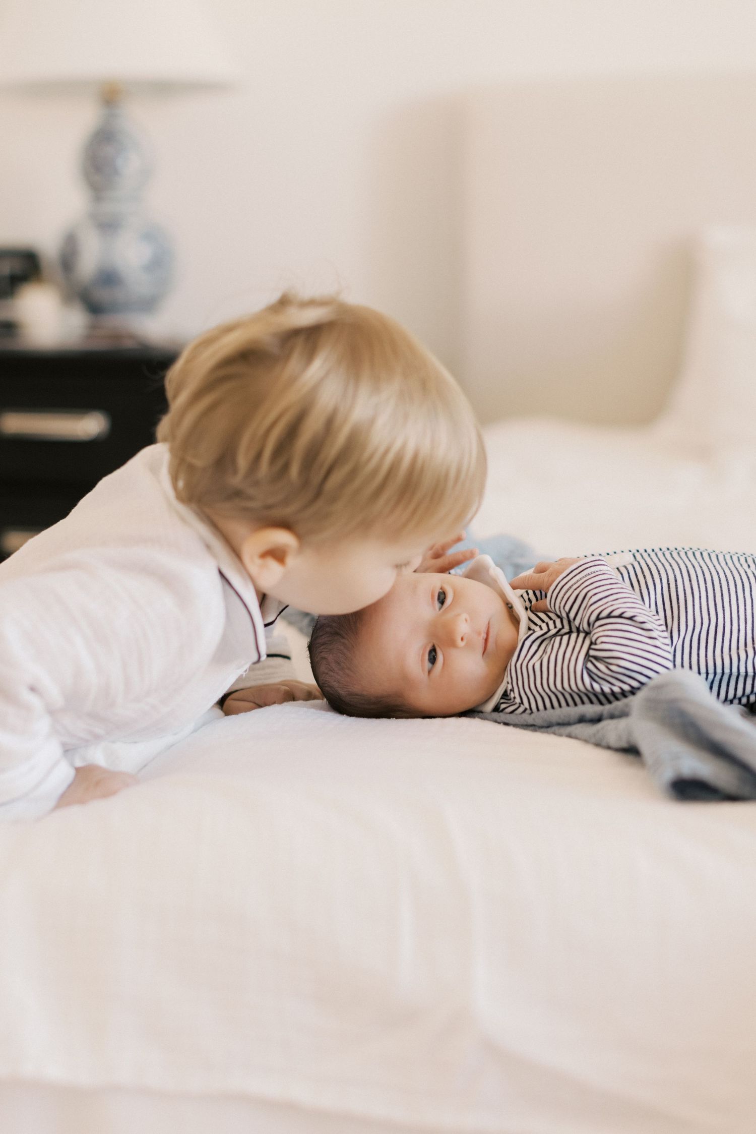 Blonde toddler gently kisses newborn sibling in striped outfit lying on a white bed.