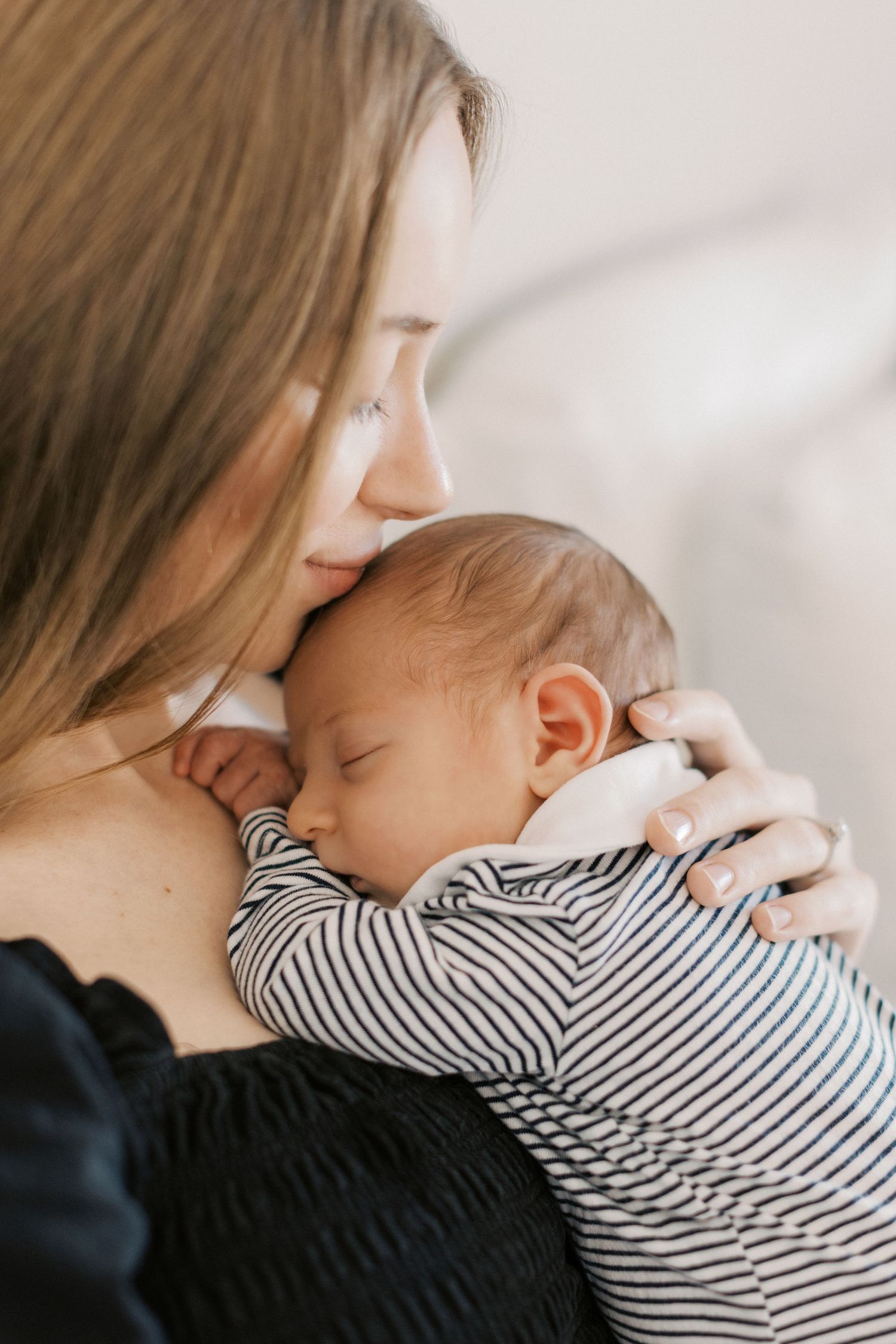 Tender moment of young mother gently nuzzling her sleeping newborn baby in striped onesie close to her chest.