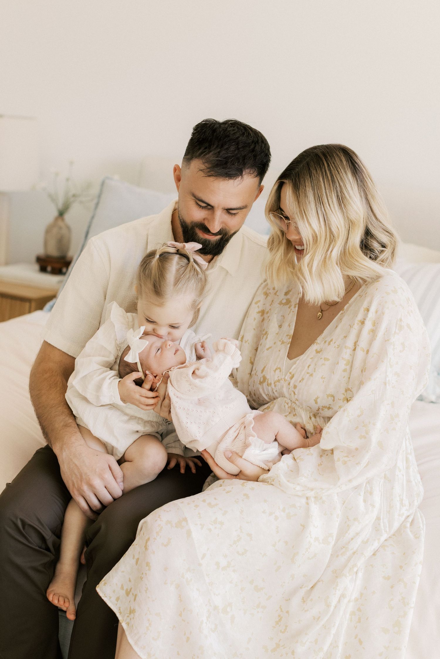 A happy family of four sits on a bed, parents smiling at their toddler and newborn baby in soft, neutral tones.