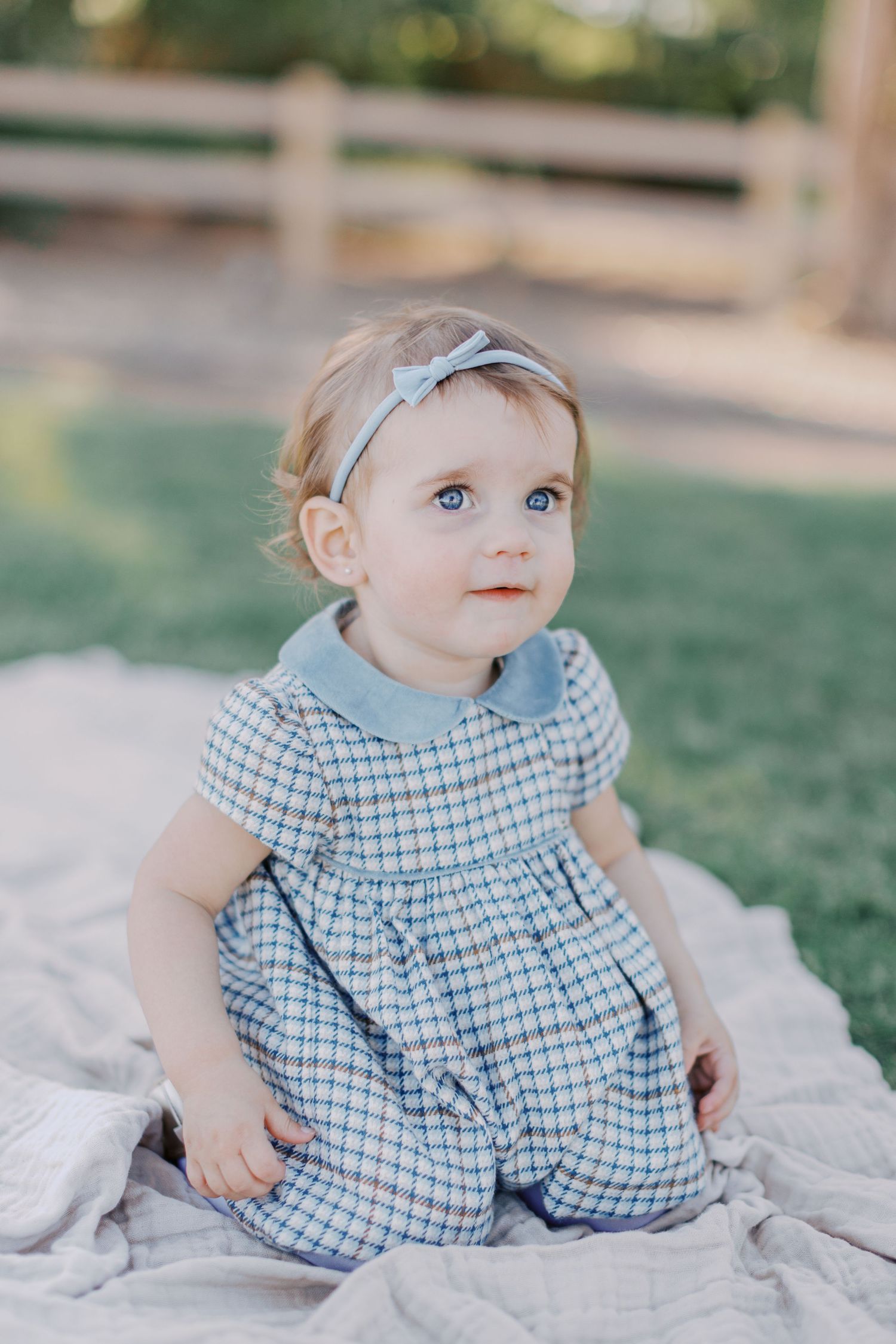 Baby girl in blue plaid dress and bow headband sits on blanket outdoors, looking up with bright blue eyes.