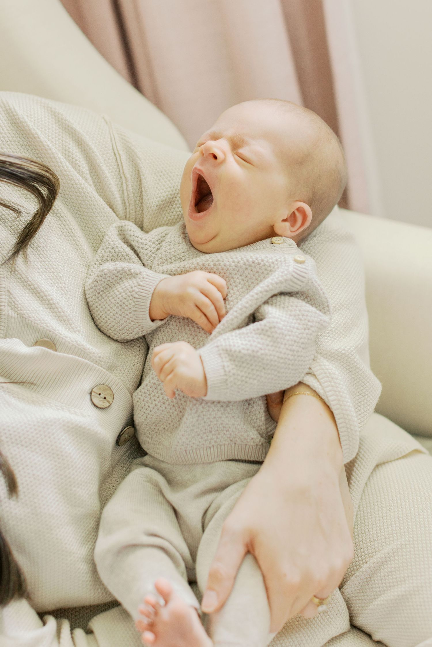Adorable newborn baby yawning while being held in mother's arms, dressed in a cozy beige knit outfit.