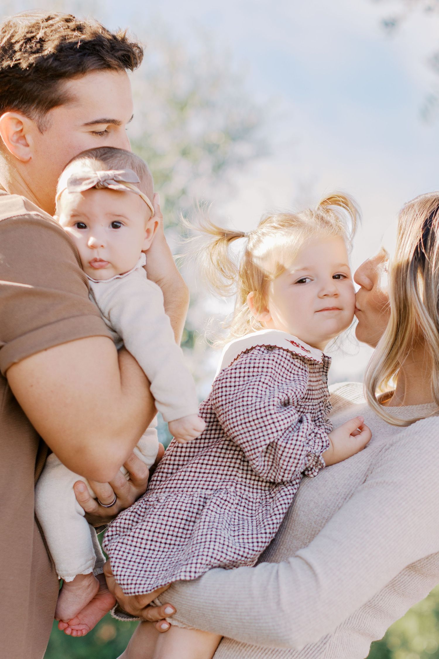 Parents holding infant and toddler daughter in plaid dress during outdoor family portrait.