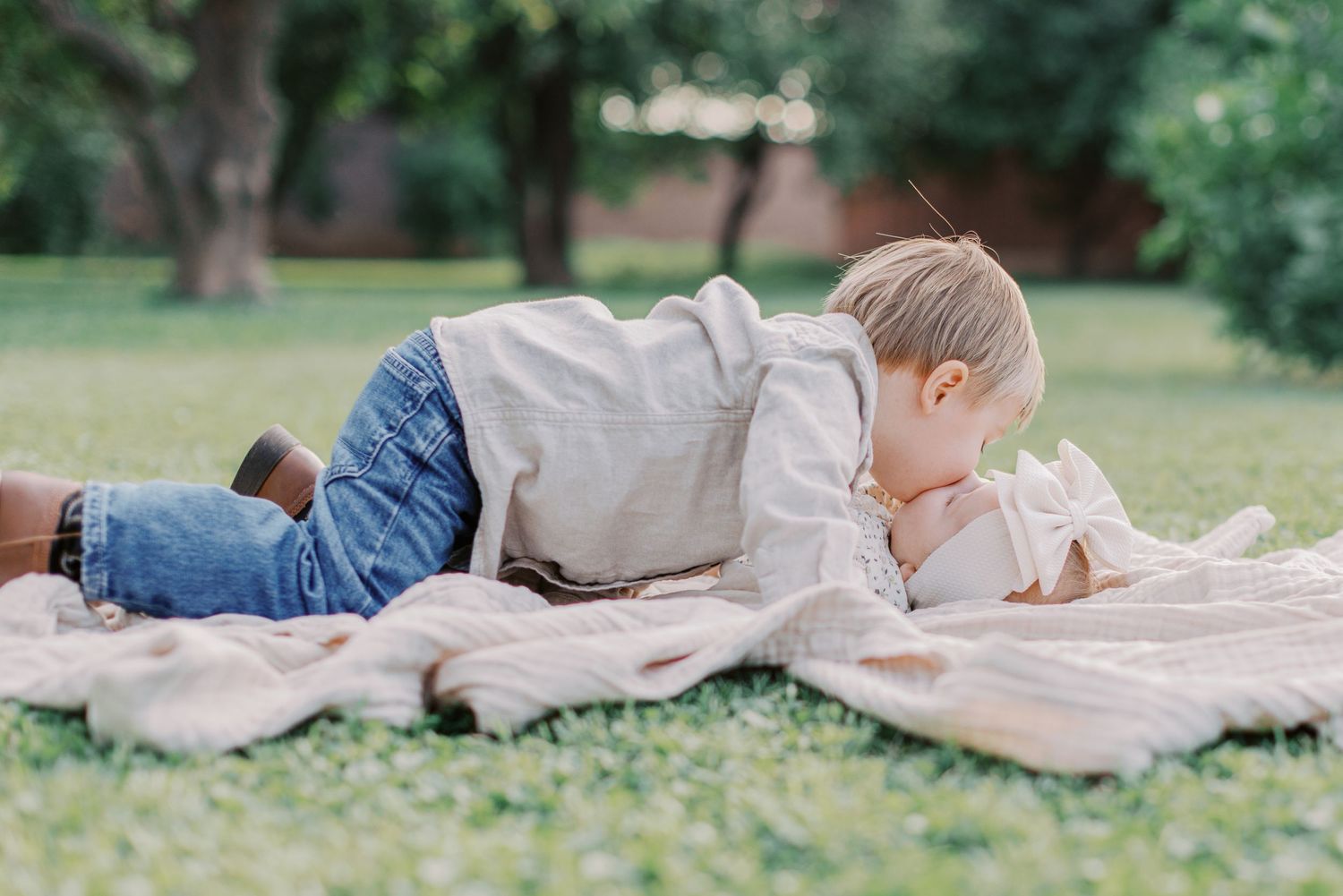 Young boy in jeans lying on a blanket in the grass, cuddling with a baby outdoors in a park.