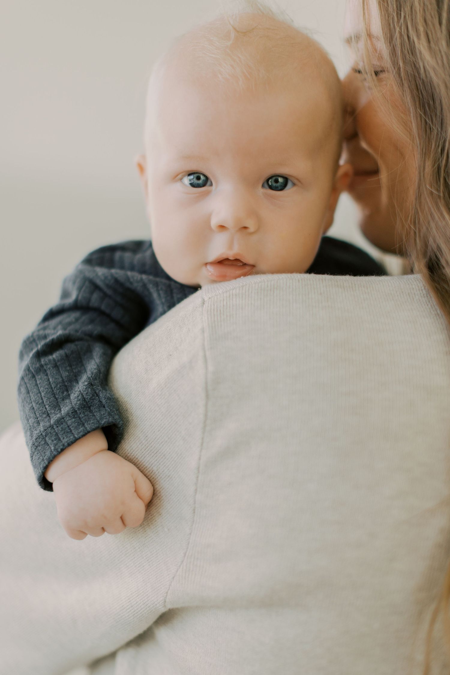 Blue-eyed baby in dark knit outfit looking over mother's shoulder during newborn session.
