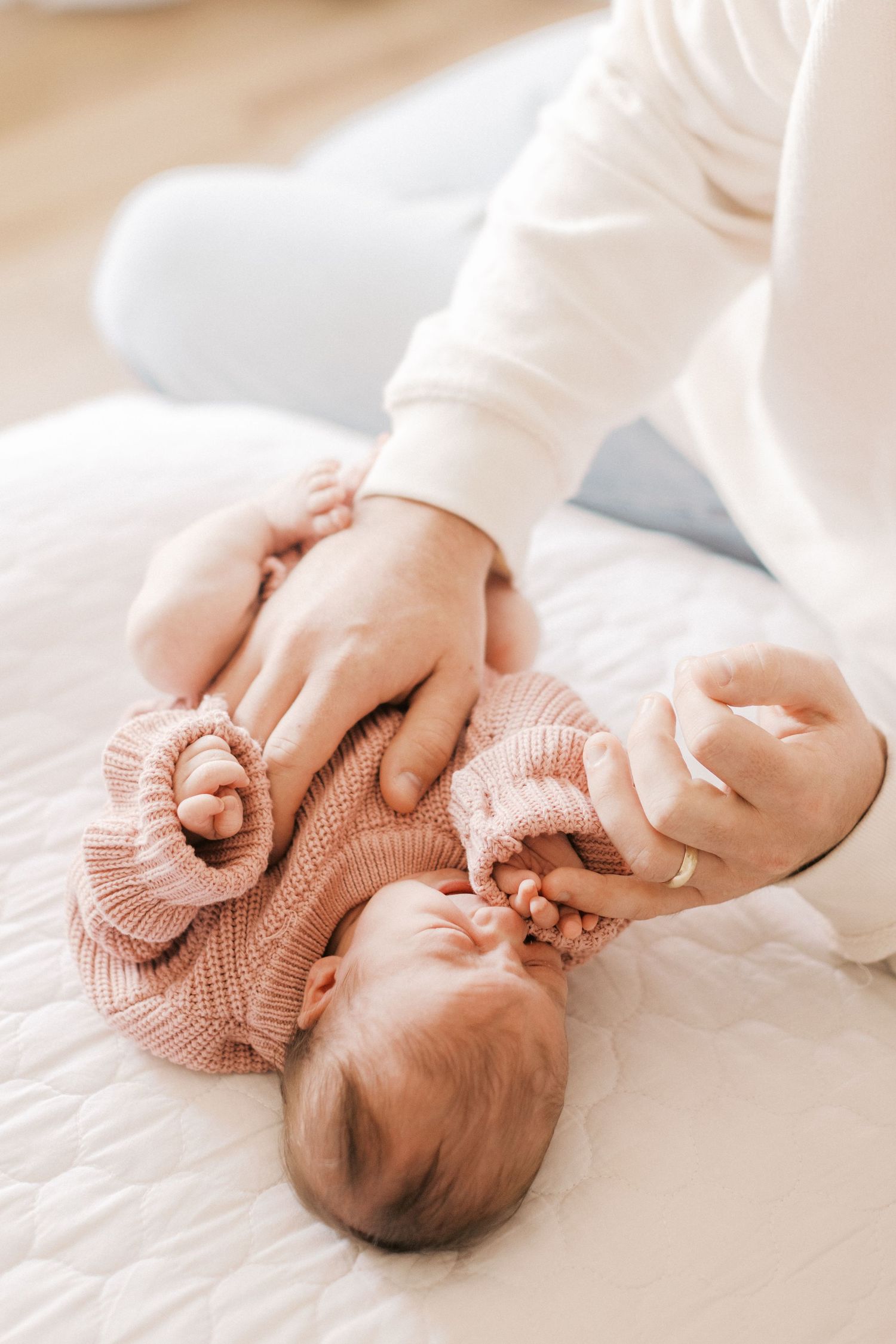 Adult hands gently holding a newborn baby in a pink knit outfit lying on a white bed.