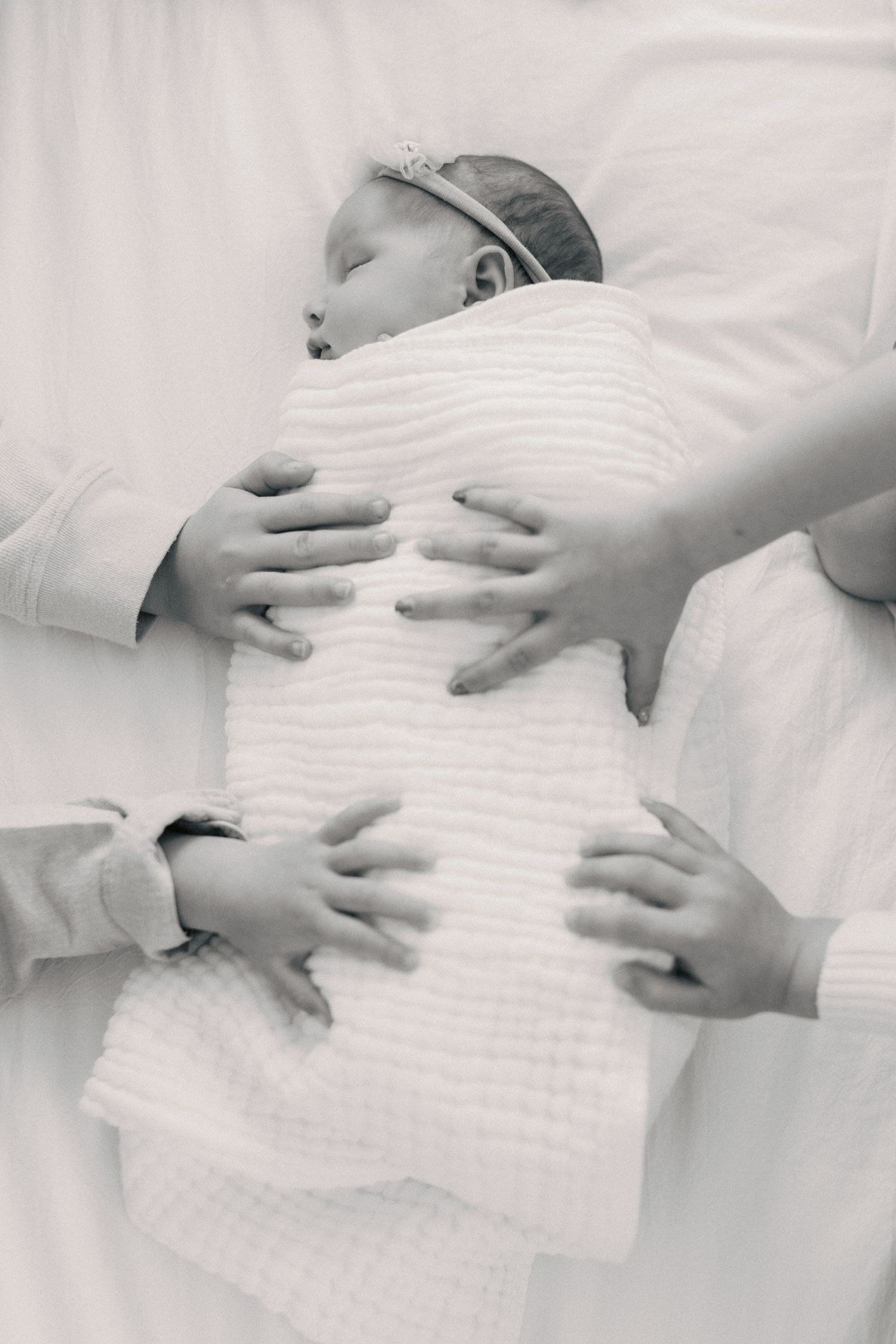 Black and white photo of sleeping newborn baby wrapped in white blanket with family hands gently resting on them.