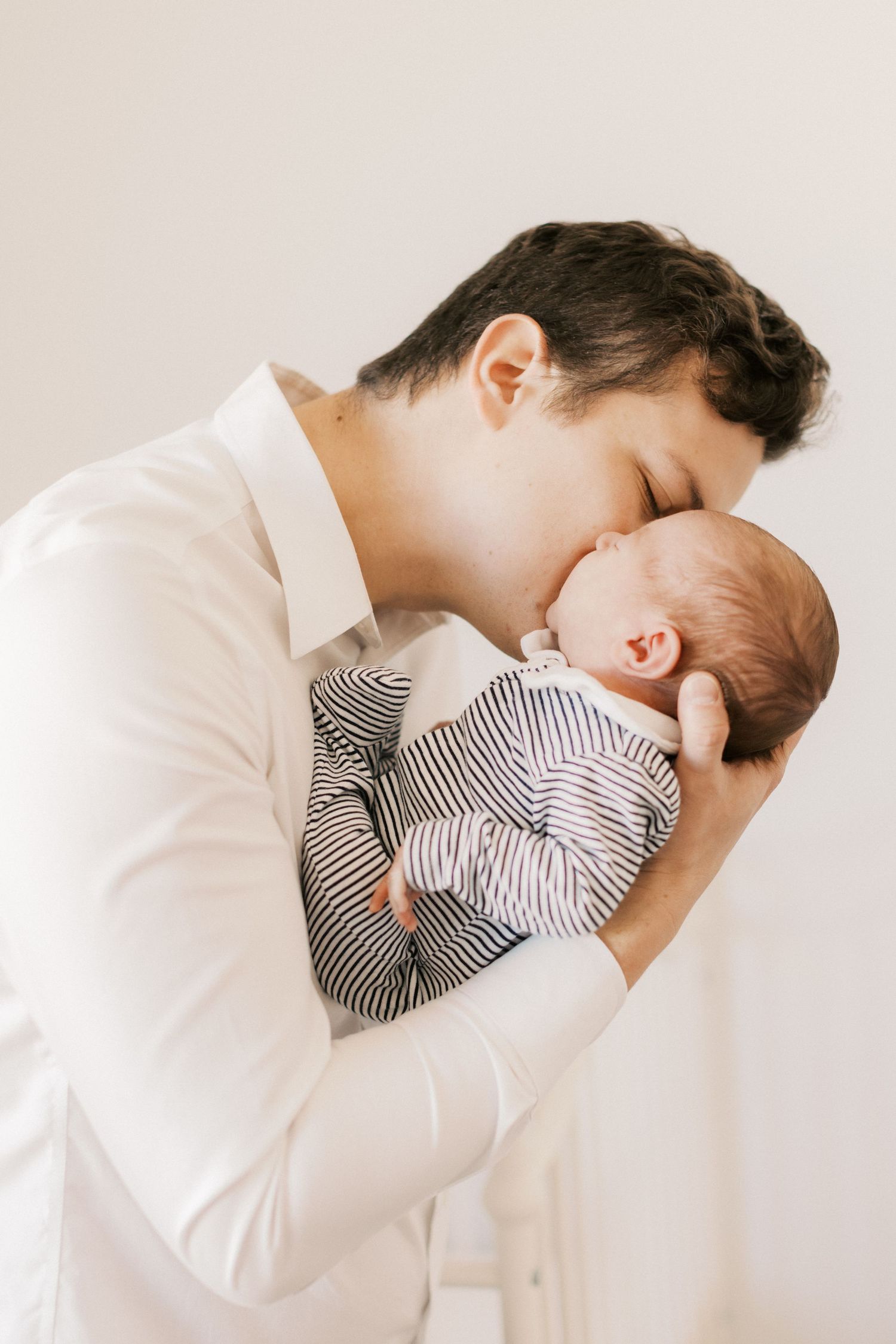 A father in a white shirt tenderly kisses his newborn baby dressed in a striped onesie.