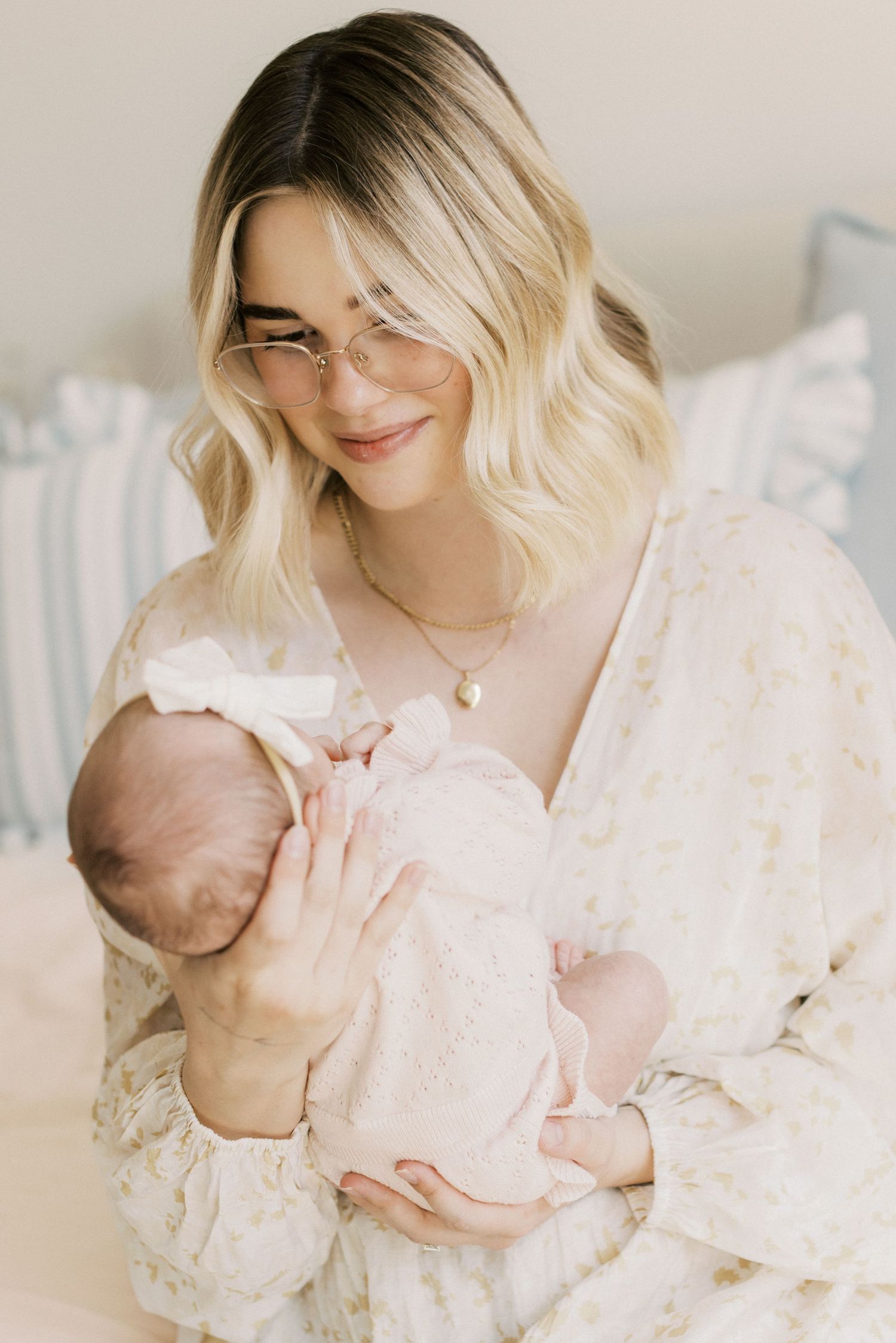 Young blonde mother with glasses tenderly holding her newborn baby in a soft floral dress.