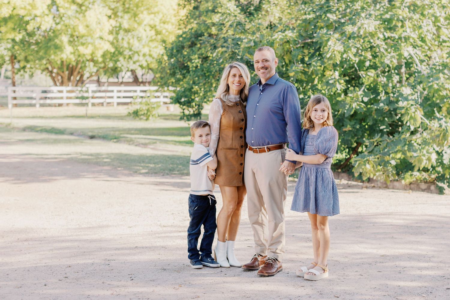 Portrait of smiling family of four with young boy and girl standing outdoors on sunny gravel path.