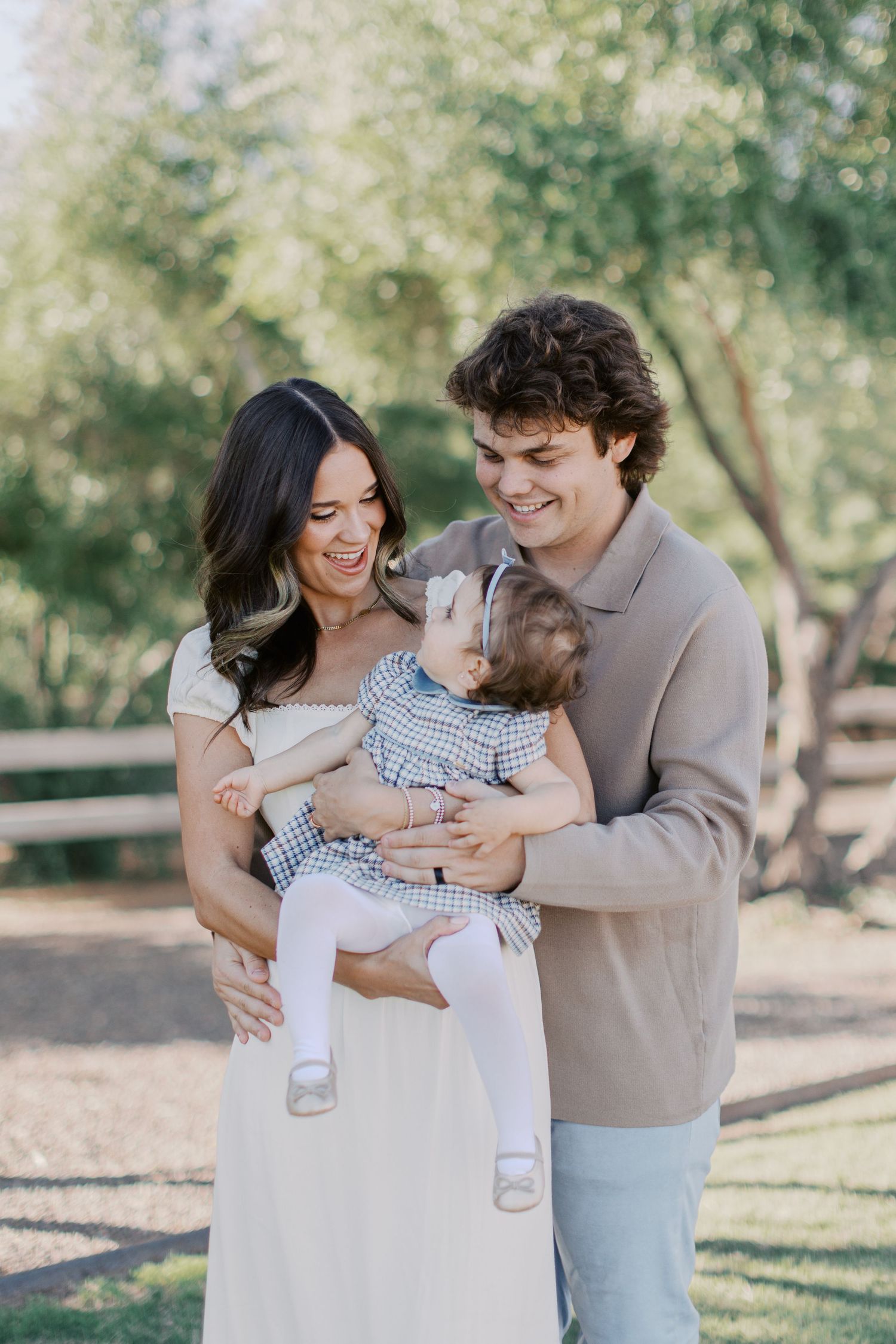 Happy young family with curly-haired man and brunette woman smiling at their baby outdoors in a sunny park.