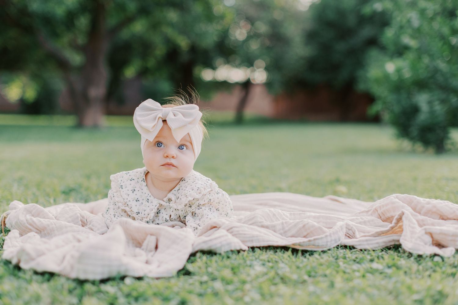 Baby girl in floral outfit and bow headband lying on a pink blanket on green grass outdoors.