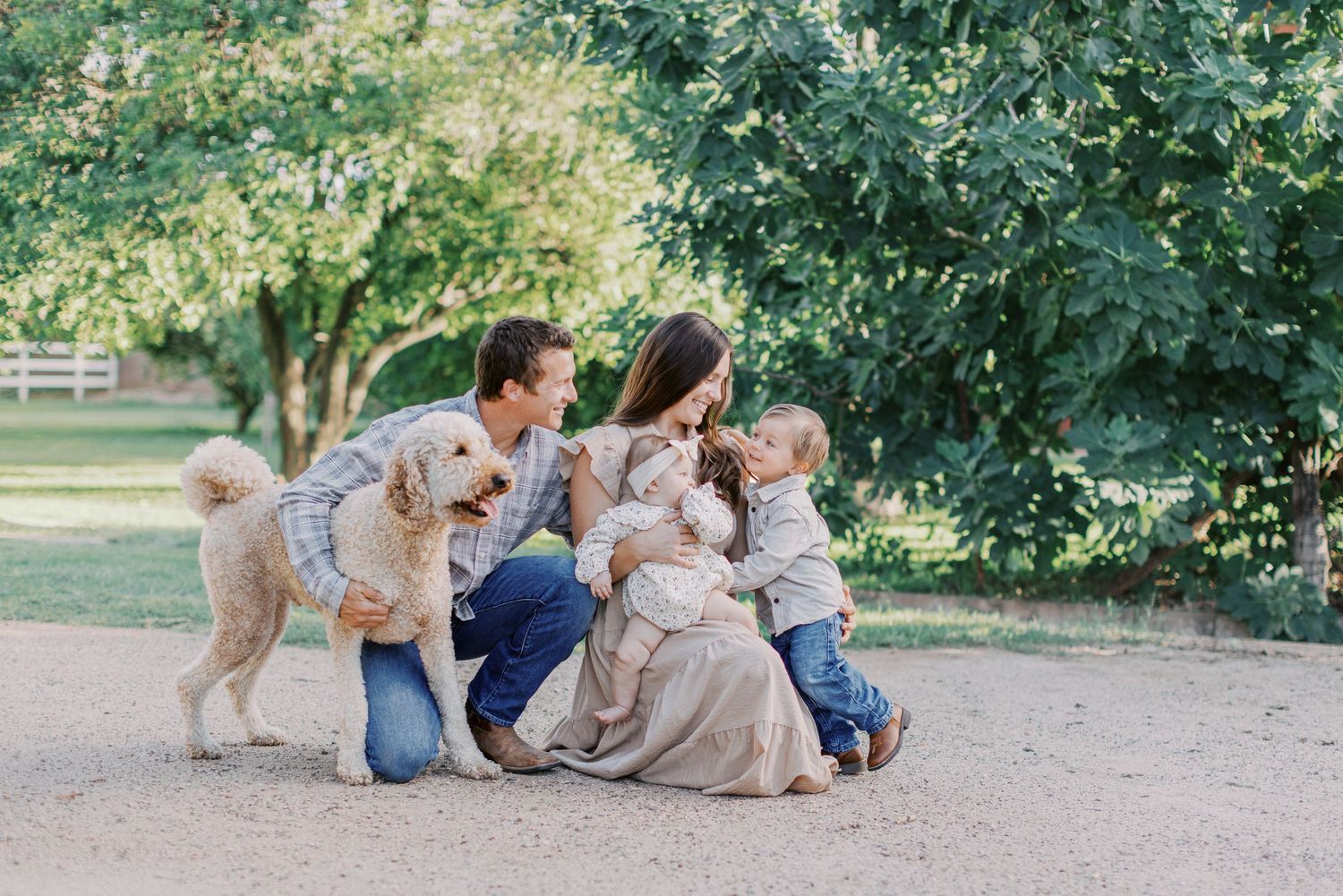 Family with toddler, baby, and golden retriever dog posing outdoors in a lush green park setting.