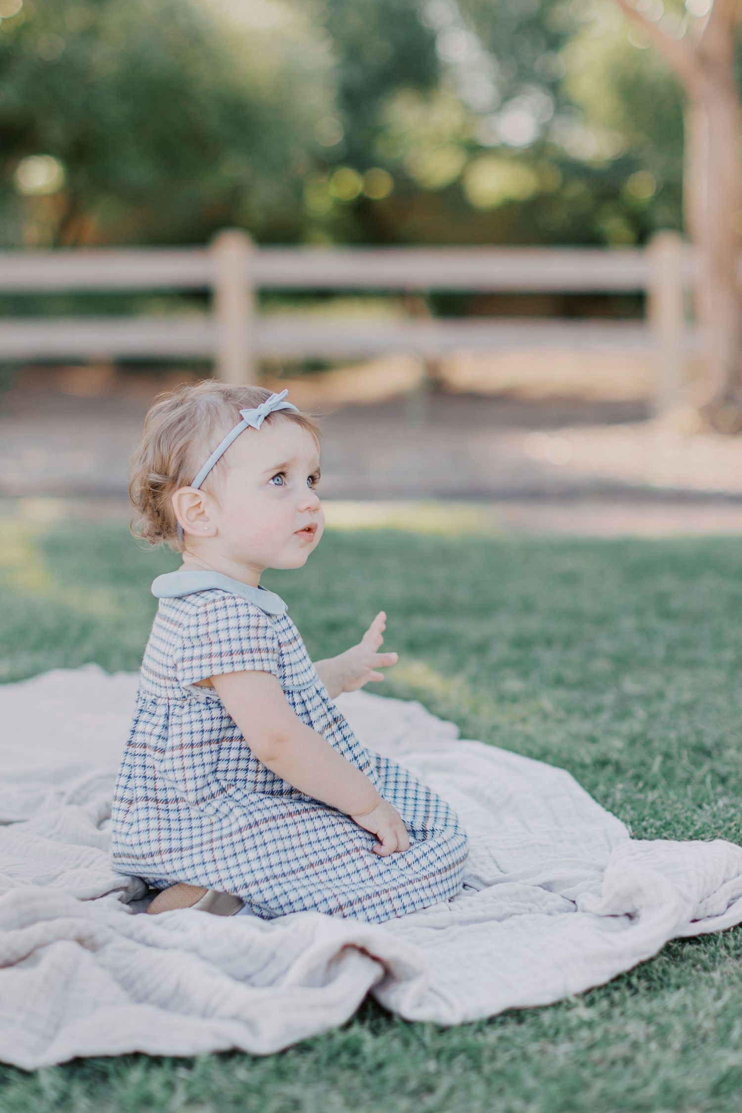 Toddler girl in plaid dress and bow headband sitting on blanket in grass outdoors.