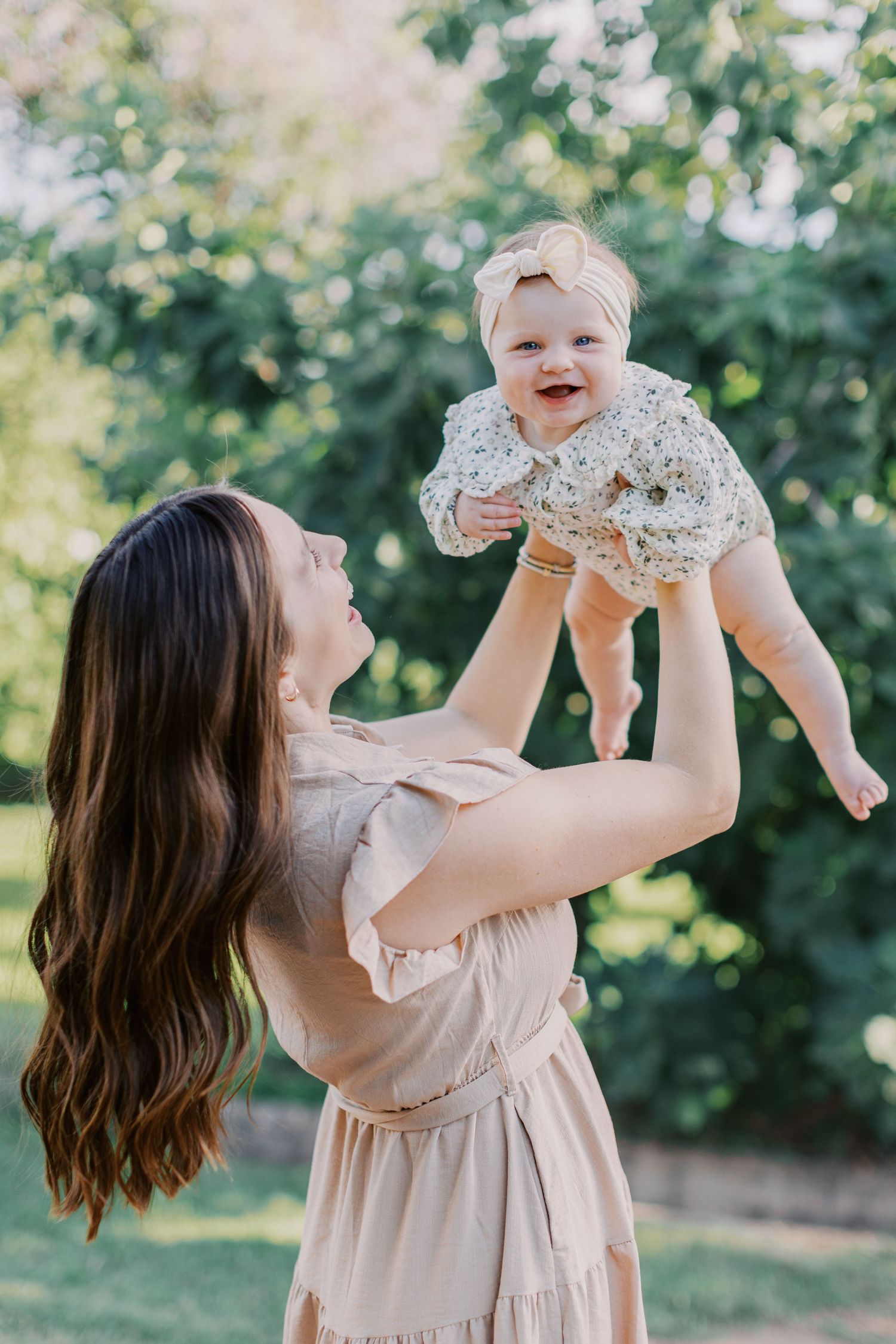 A joyful mother lifts her smiling baby girl in a sunny green park, both wearing soft floral and neutral dresses.