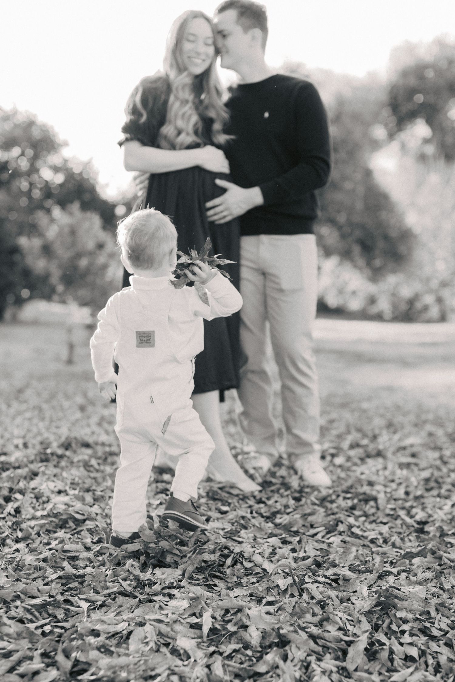 Black and white family photo of couple embracing in park while toddler in white outfit walks toward them on fallen leaves.