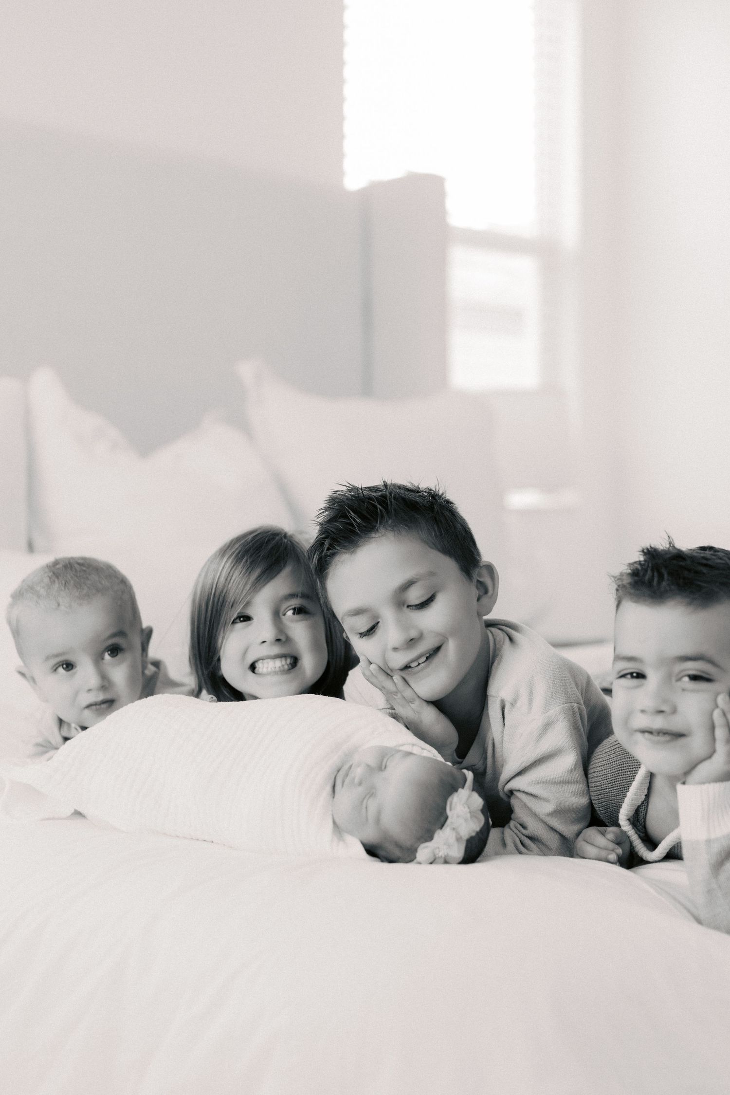 Black and white photo of four young siblings lying on a white bed together with a newborn baby.