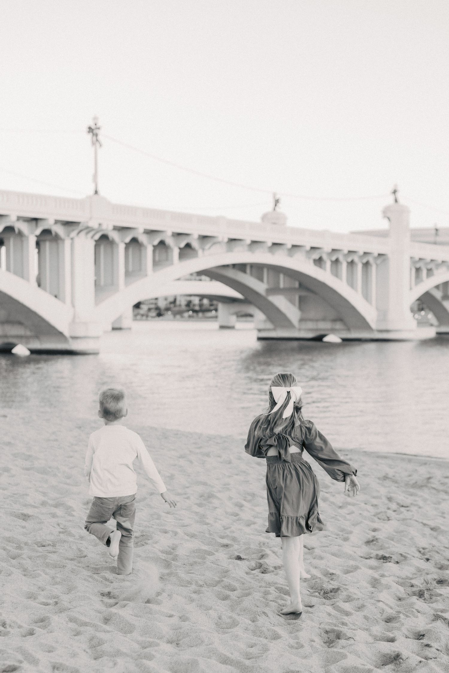Two children running on a sandy beach toward an arched bridge over a river in a black and white photo.