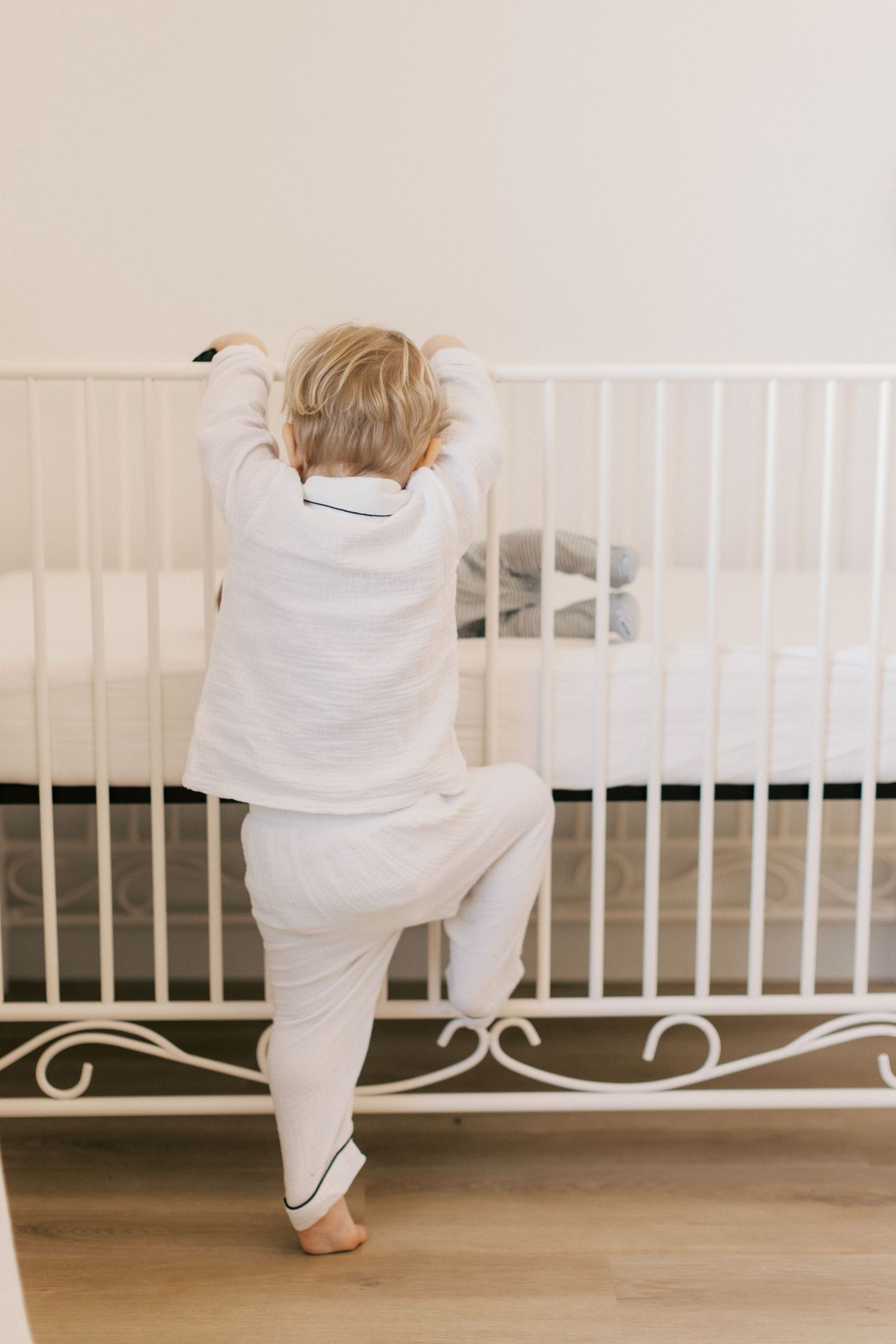 A toddler in white pajamas stands on tiptoes at a white metal crib, reaching over the rail in a cozy bedroom.