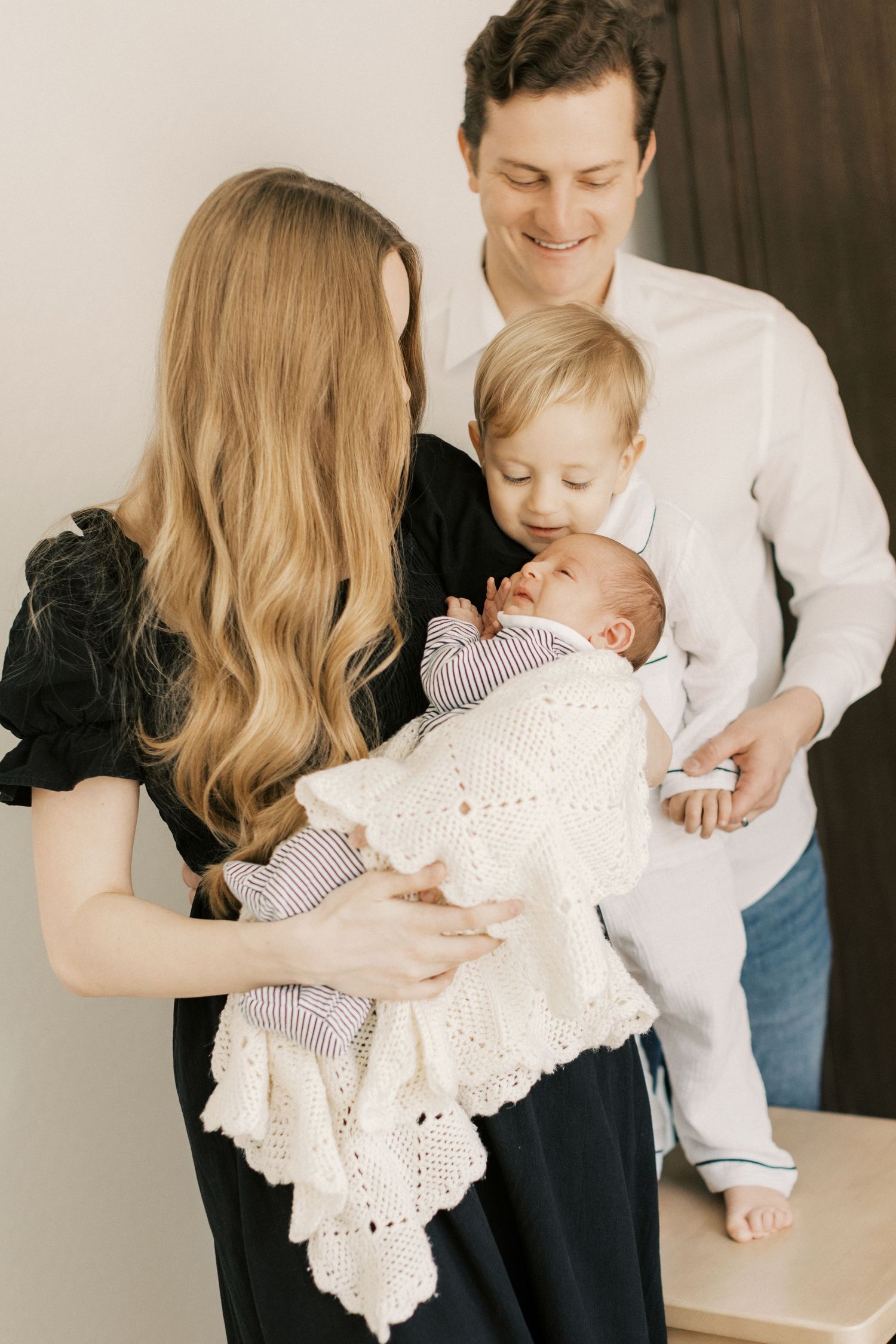 Happy family with newborn baby wrapped in white knit blanket, parents smiling with toddler sibling indoors.