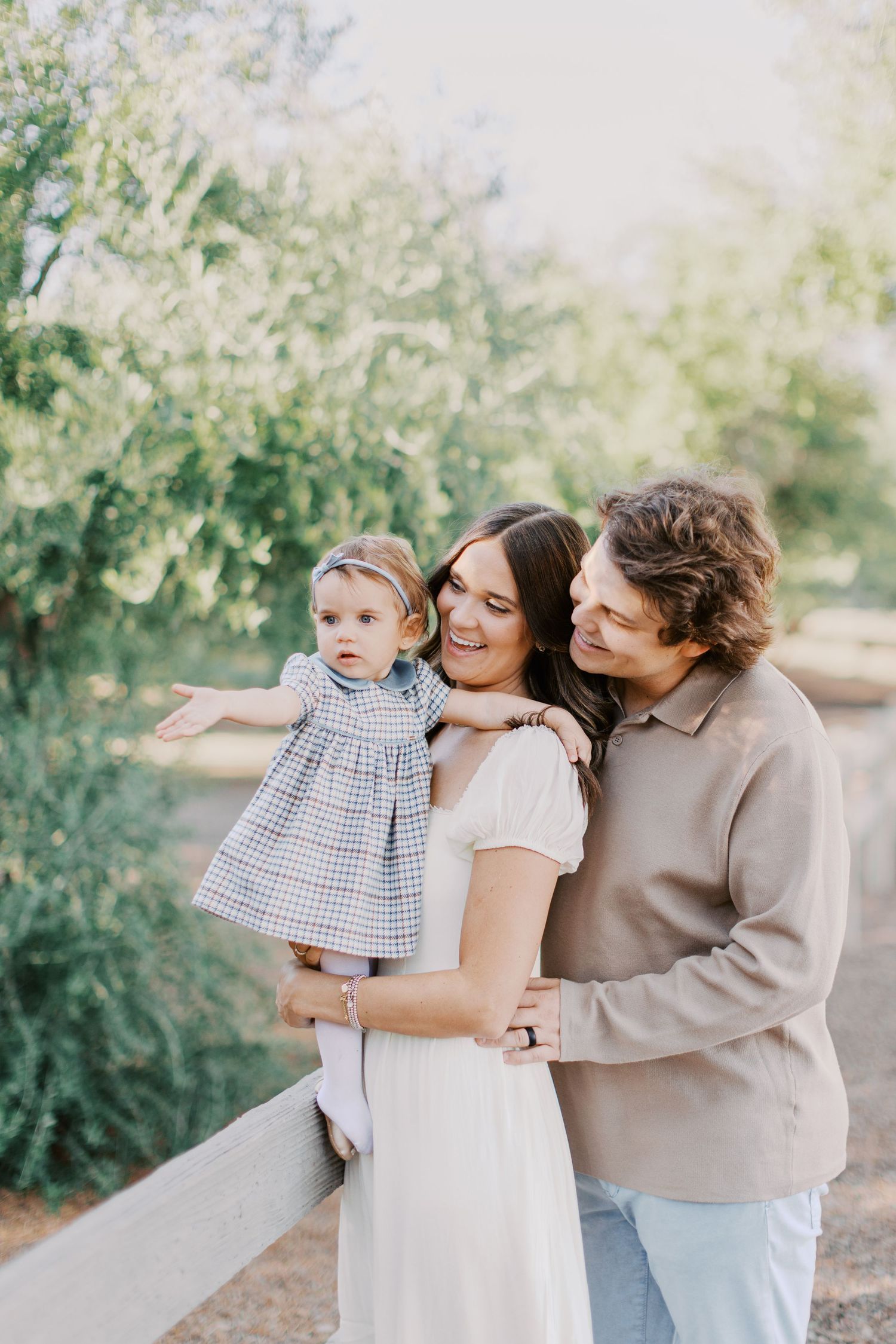 Happy young family with toddler daughter posing by white fence surrounded by lush green trees outdoors.