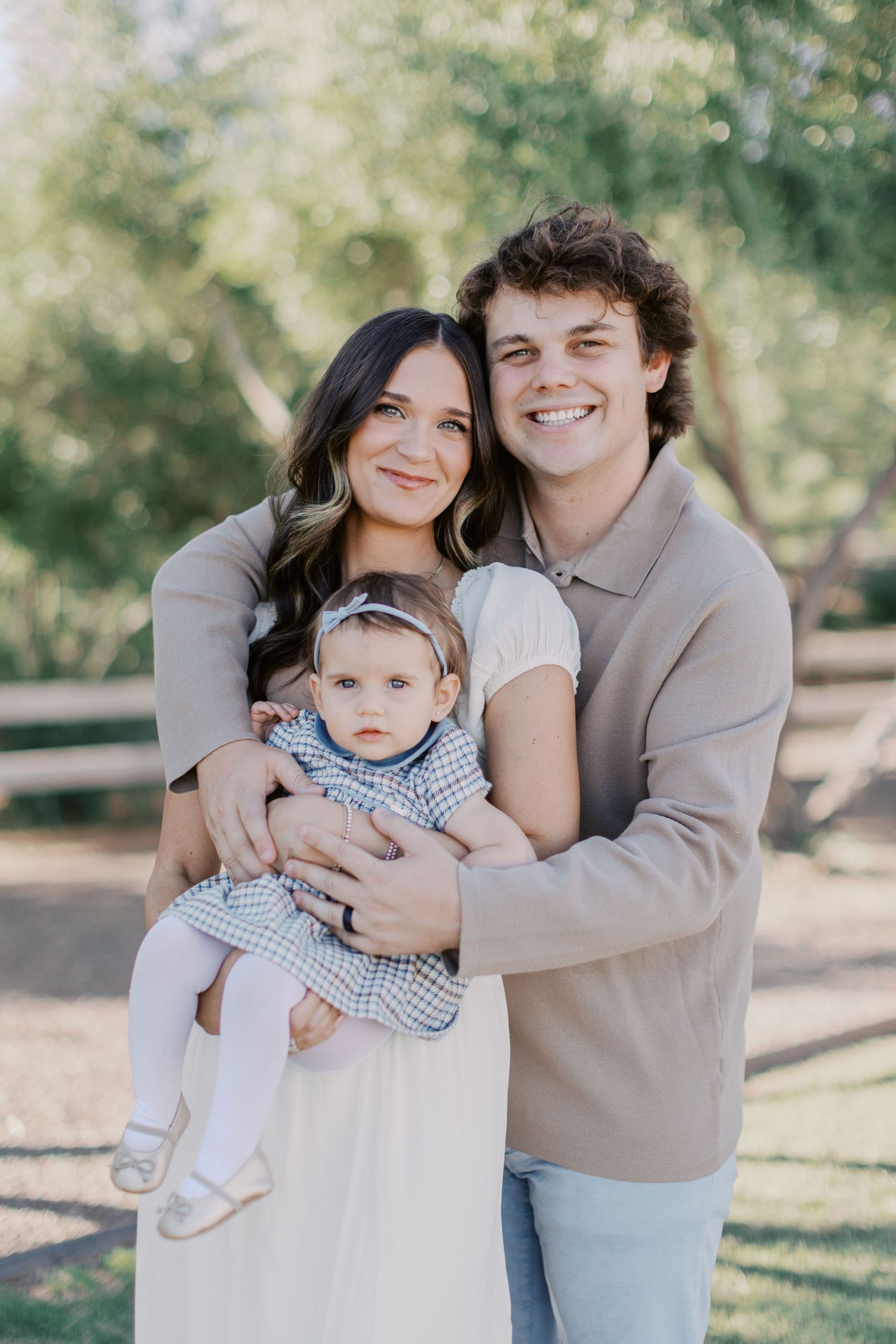 Young couple smiling outdoors holding their baby girl in a blue dress, posing for a family portrait in a park.