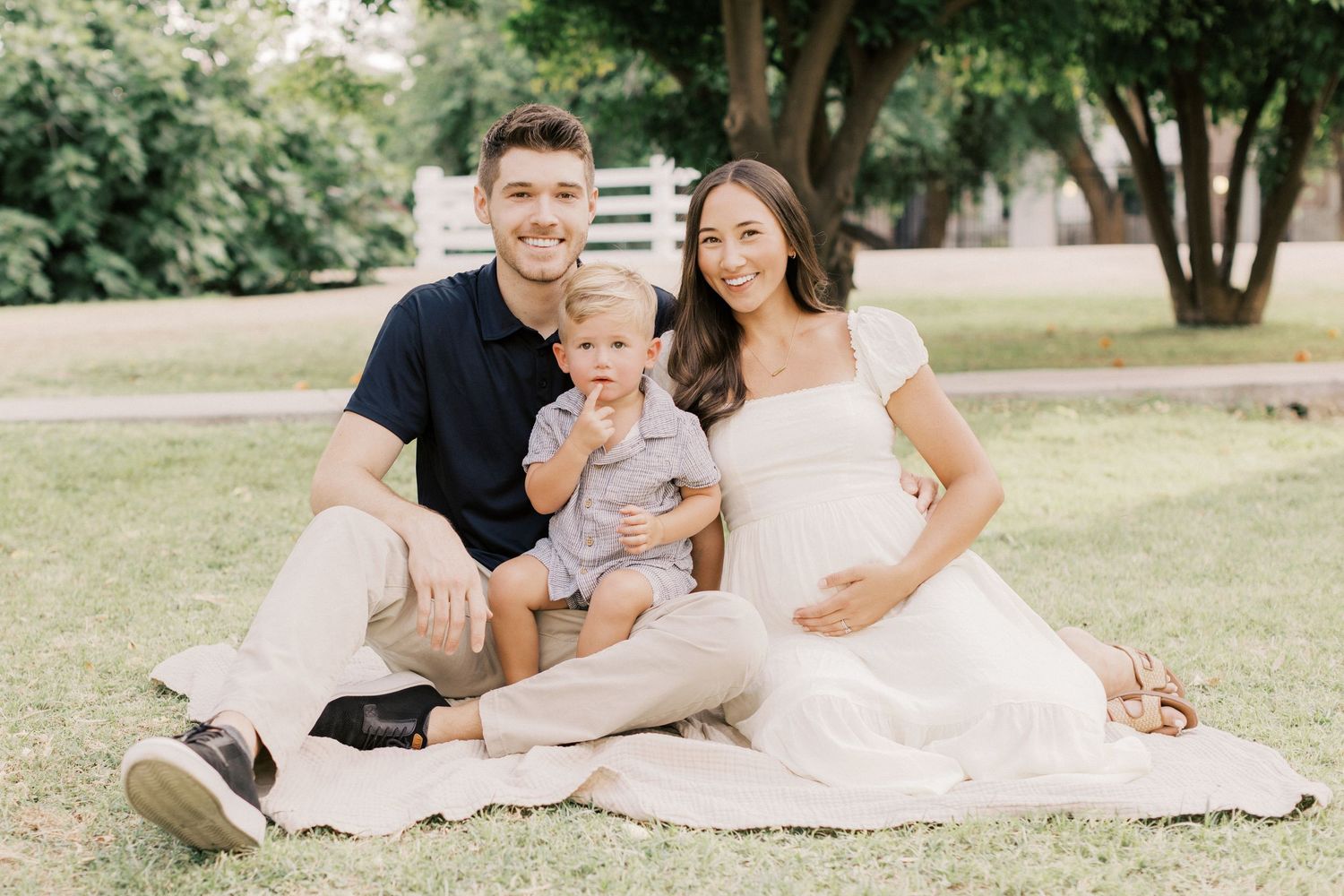 Smiling family with pregnant mother and toddler sitting on blanket in sunny green park.