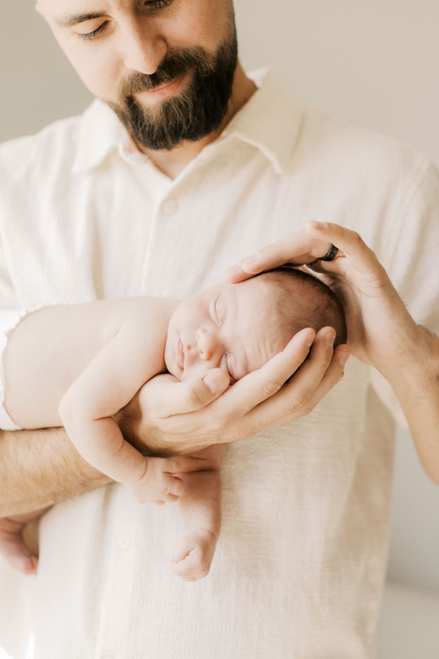 Bearded father in white shirt tenderly holds sleeping newborn baby in his hands, soft natural light.