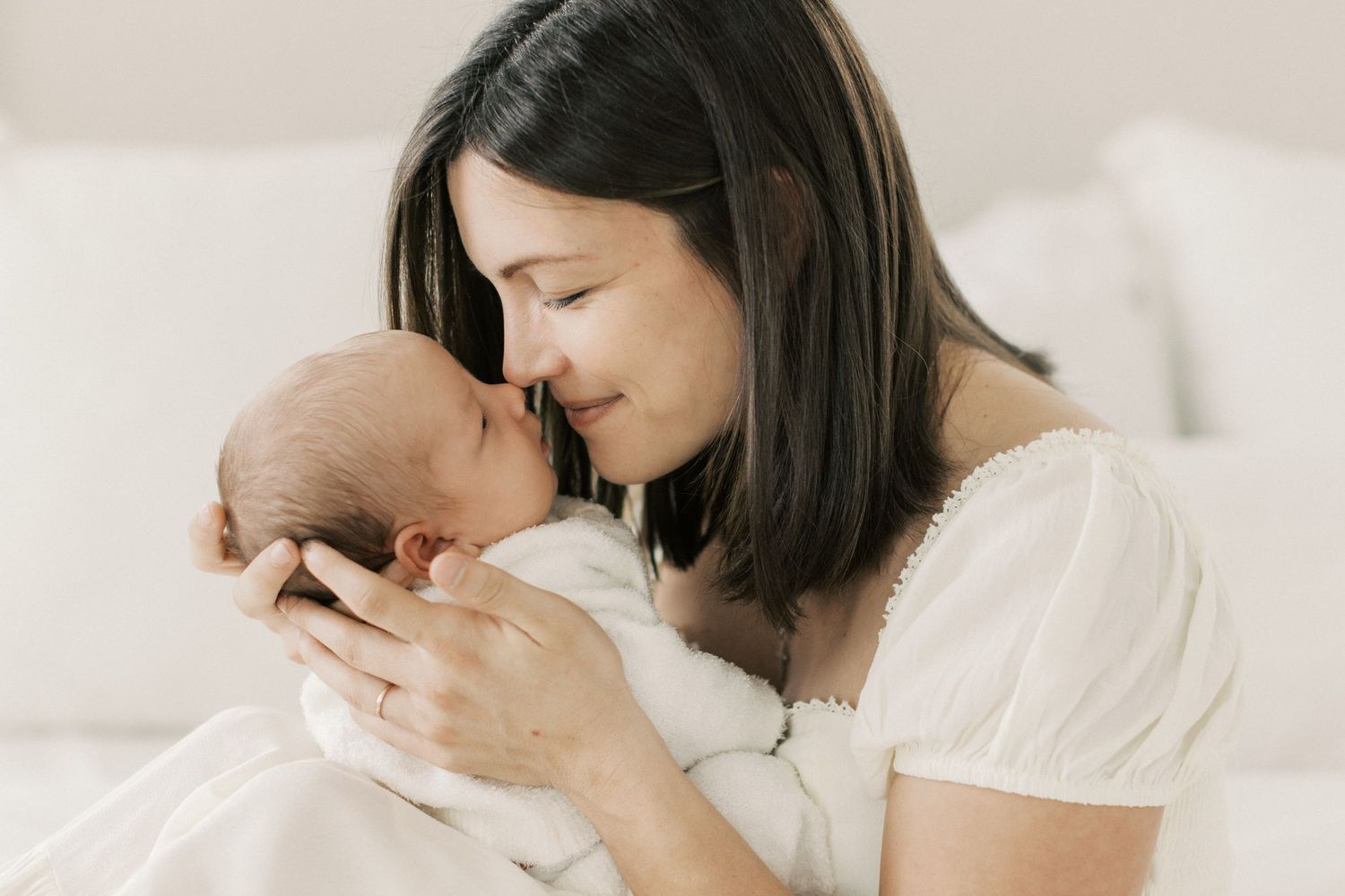 A dark-haired mother in white tenderly holds her newborn baby close, touching foreheads gently.