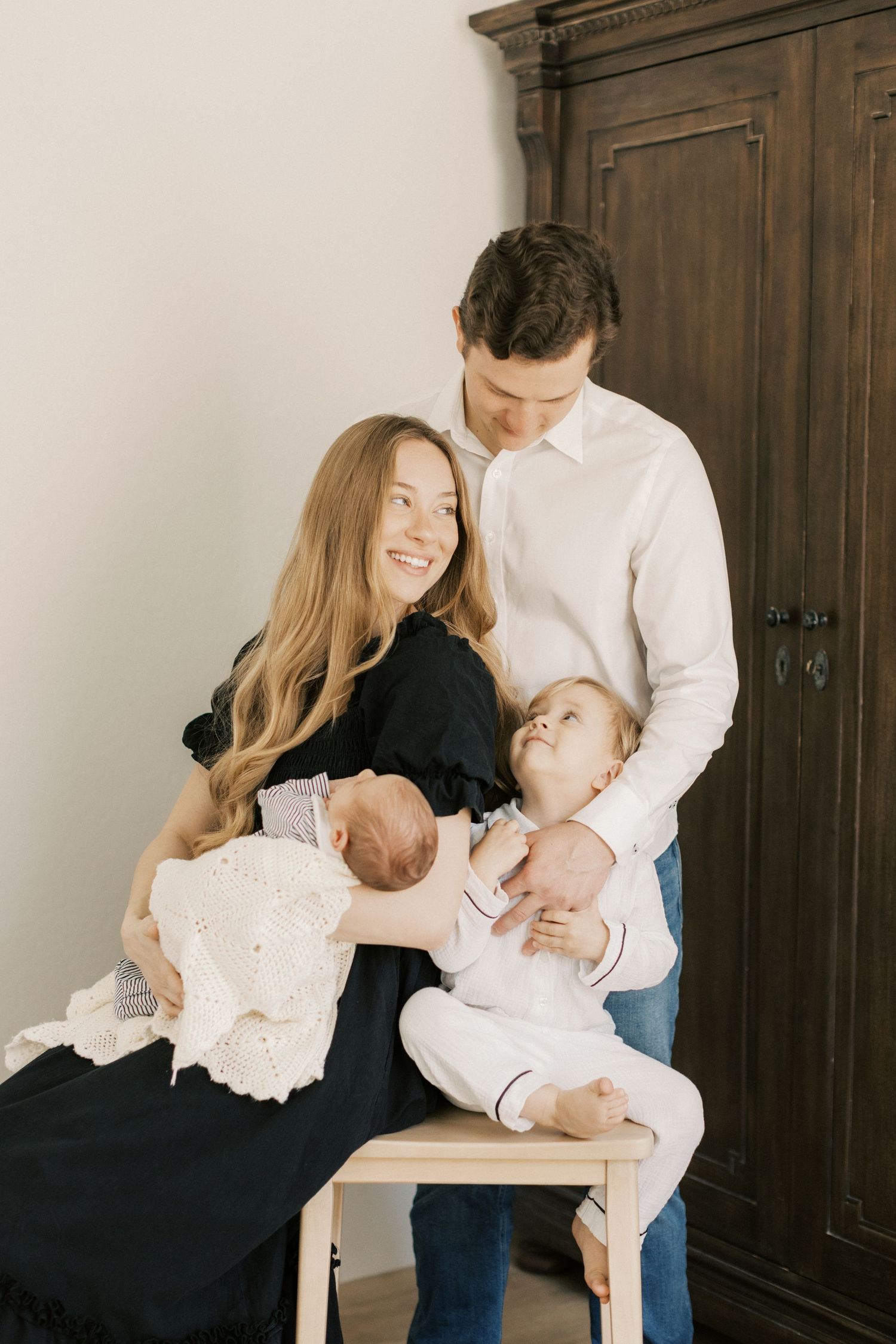 Smiling mother in black dress holds newborn while toddler sits on stool beside father near dark armoire.