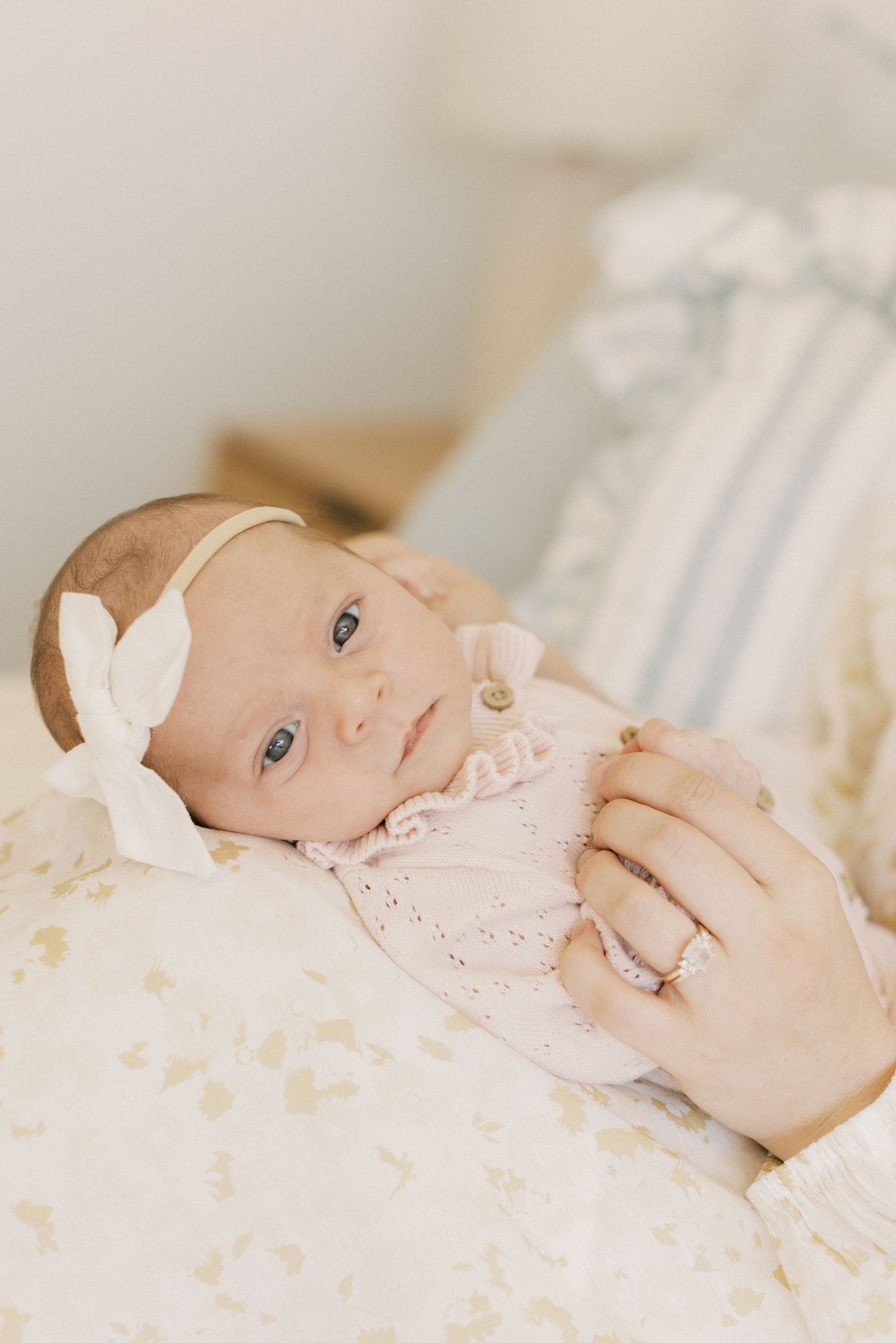 Newborn baby girl in pink knit outfit and white bow headband being cradled by mother wearing diamond ring.