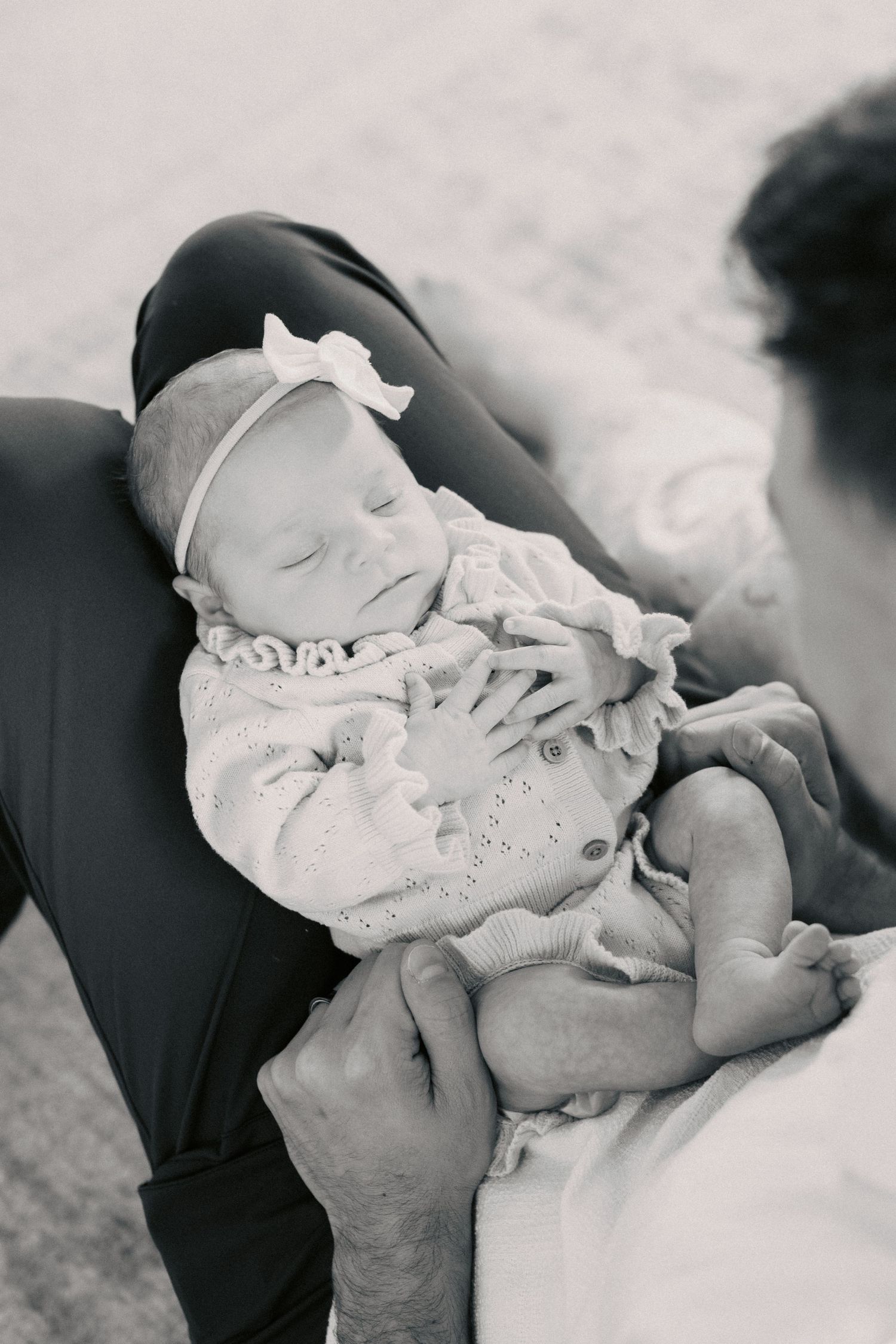 Black and white photo of a sleeping newborn baby in a white knit outfit and bow headband, cradled in fathers arms.