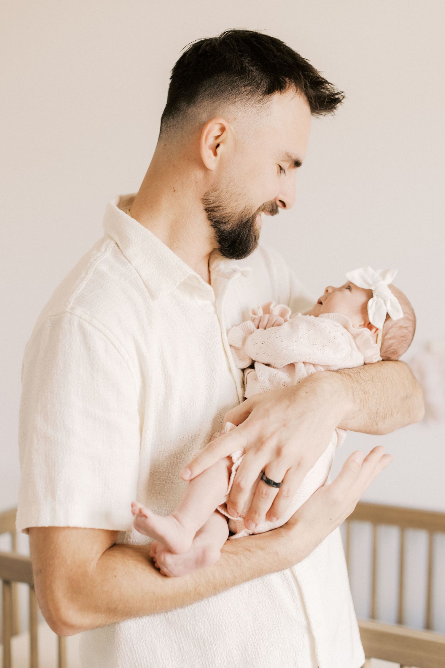 Bearded father in white shirt tenderly holds his newborn baby girl wearing a white bow headband.