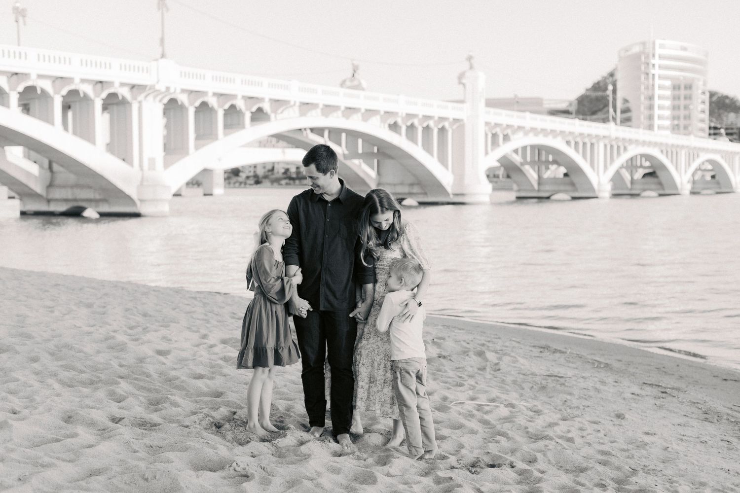 Black and white photo of a father with three young daughters standing on a sandy riverbank by a bridge.