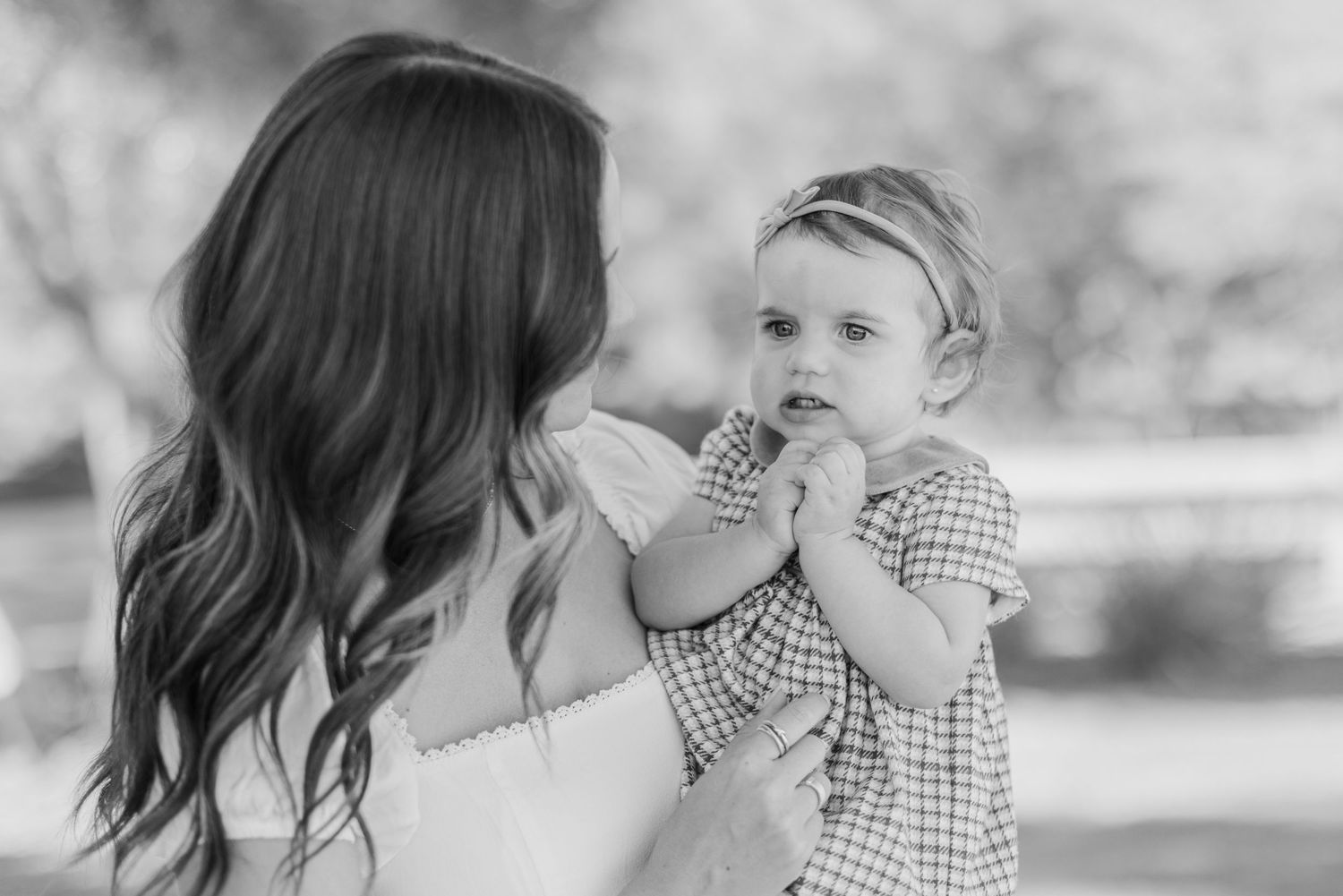 Black and white photo of a mother holding her baby girl in a checkered dress outdoors.