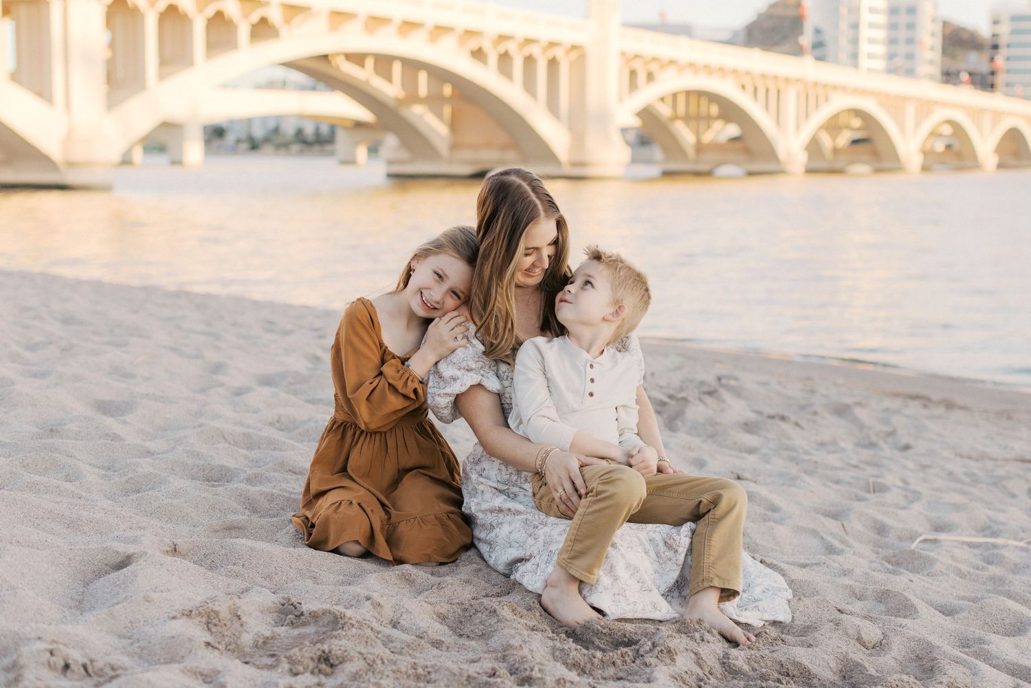Mother sitting on sandy beach with two young children hugging and smiling together.